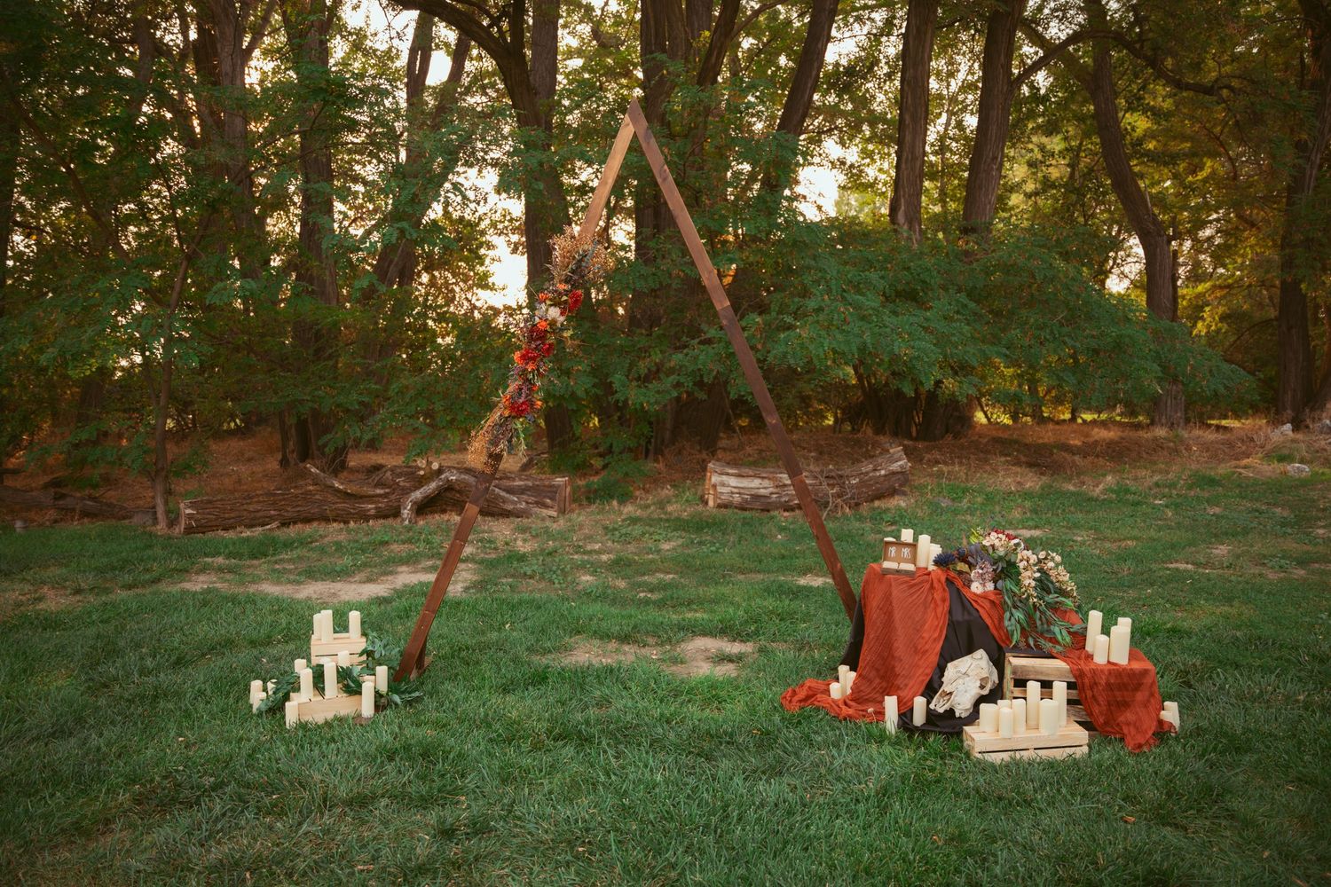 A wooden triangle arch decorated with flowers and surrounded by candles in an outdoor wedding setting.