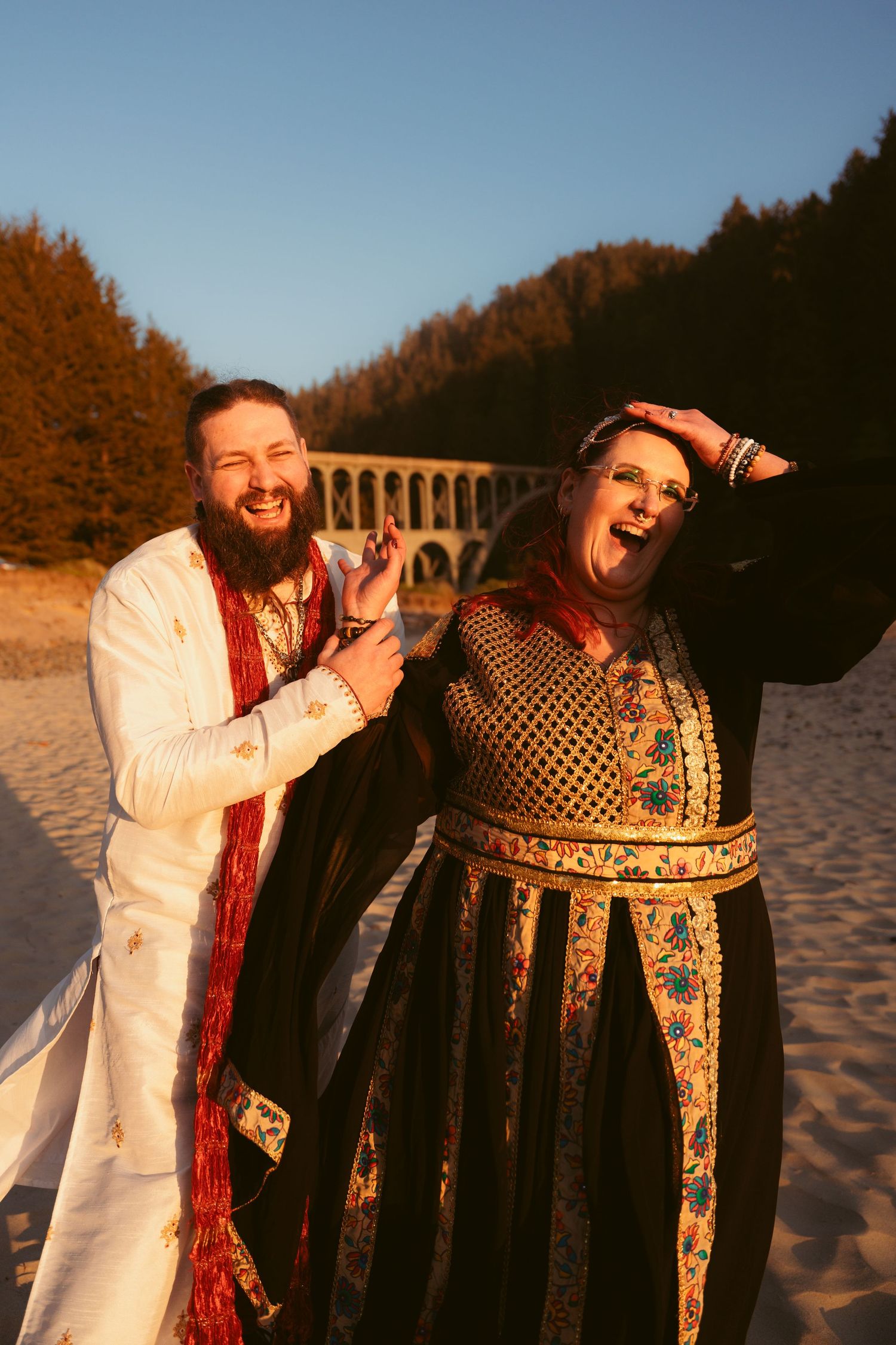 Two people in period costumes laugh together on a sandy beach during golden hour.