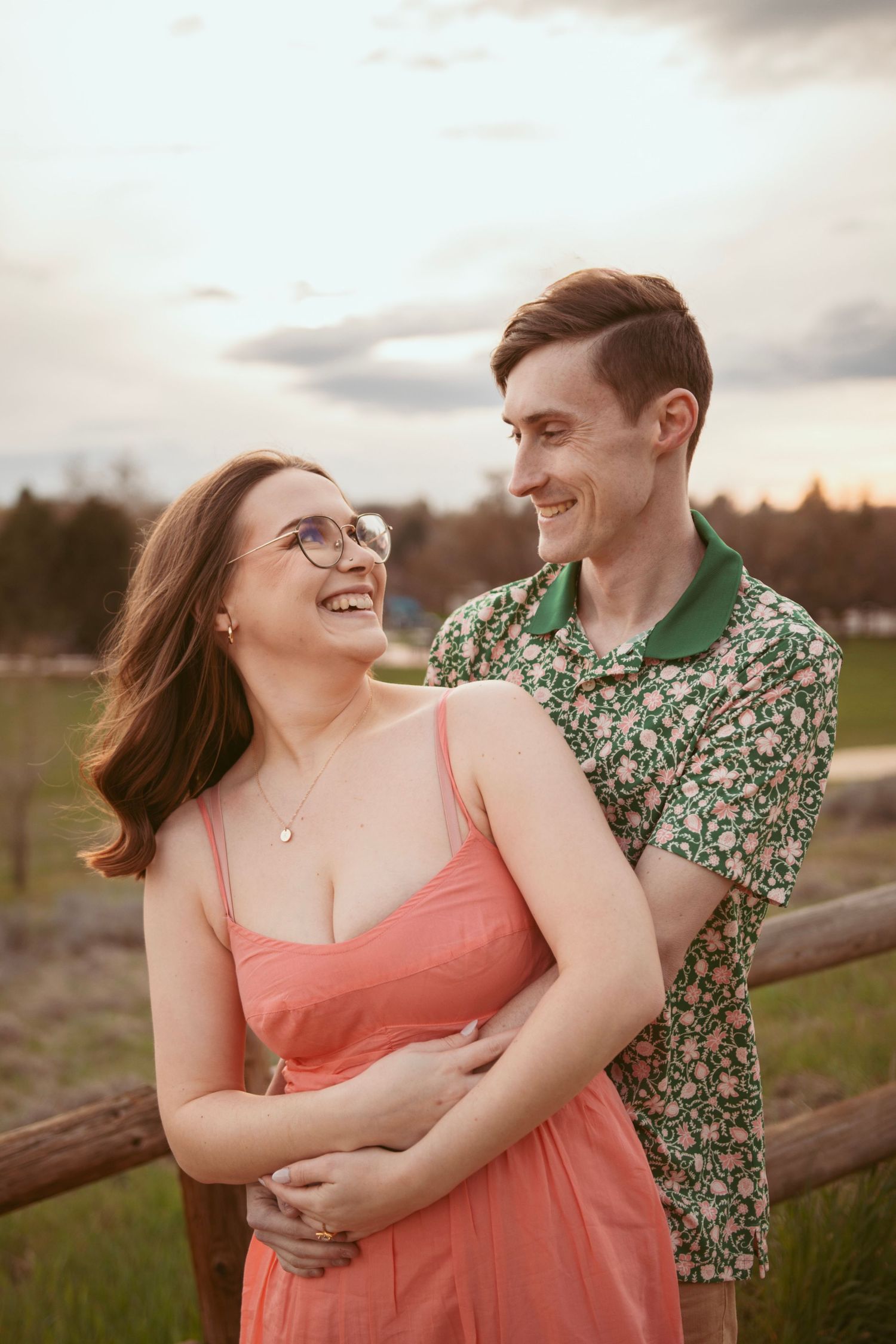 Couple shares a tender moment by rustic fence at golden hour, her in coral dress and glasses.