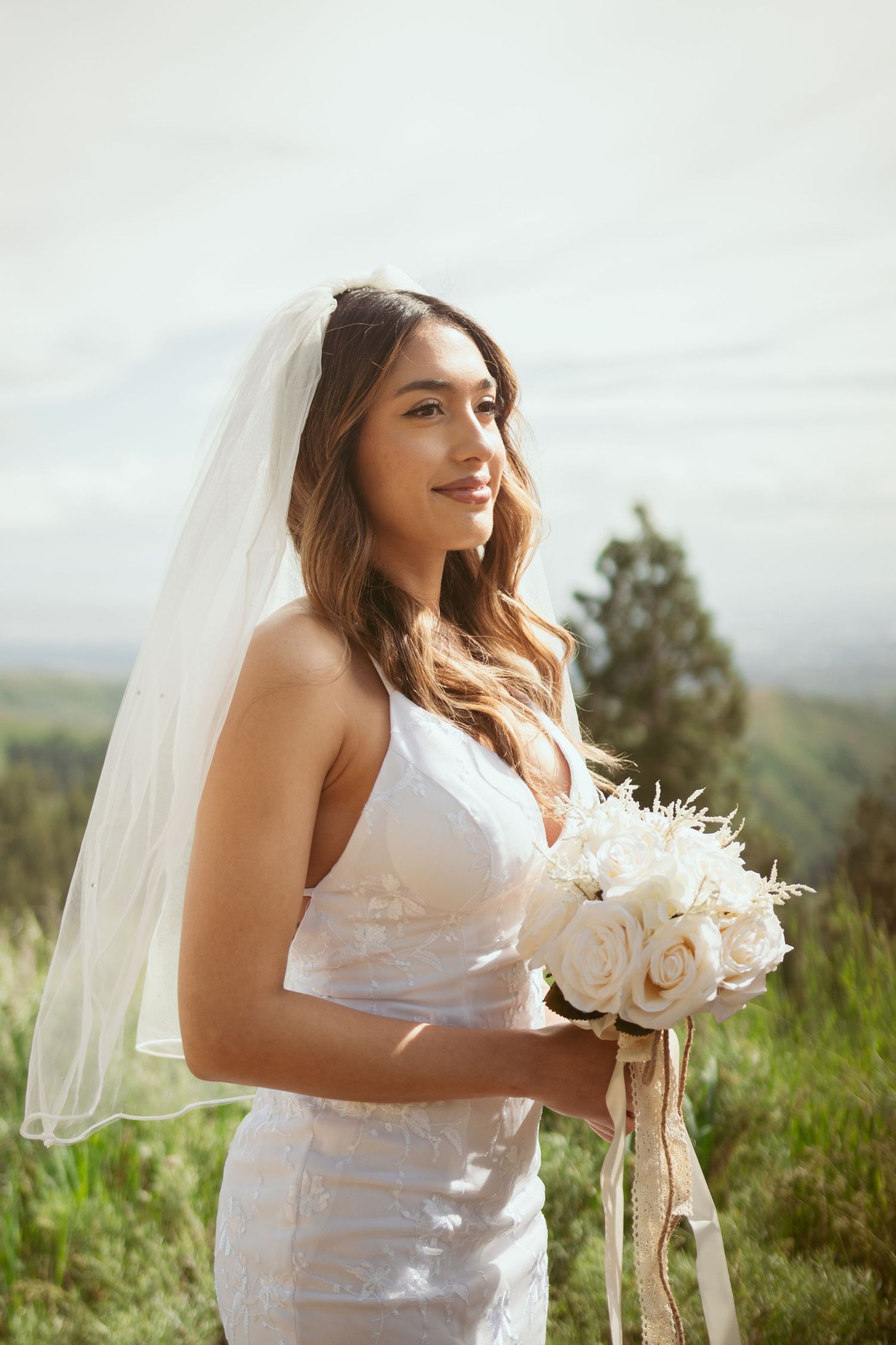 Bride in white lace dress holding elegant white rose bouquet on hilltop.