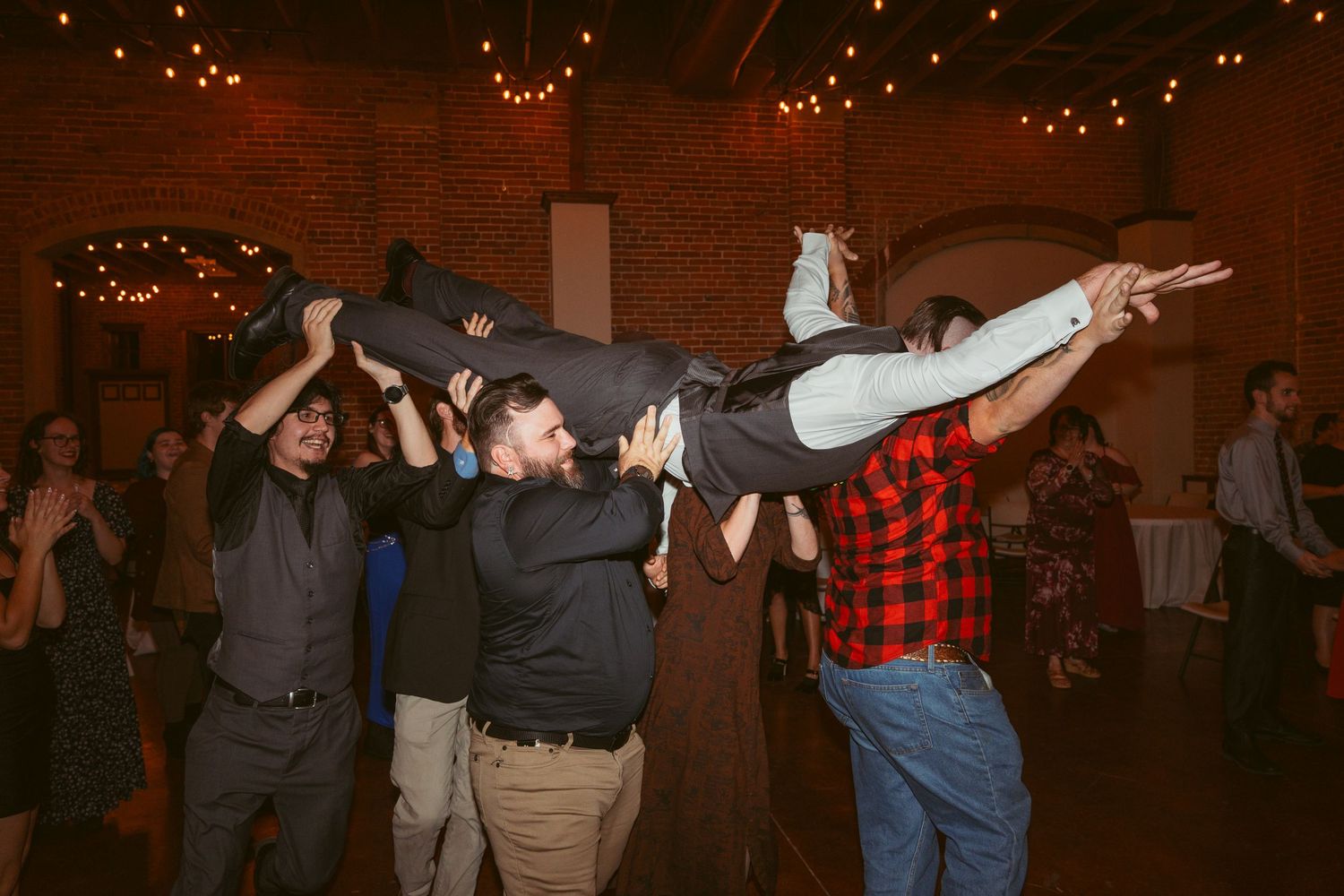 People dancing and lifting someone horizontally in the air at a party venue with string lights and brick walls.