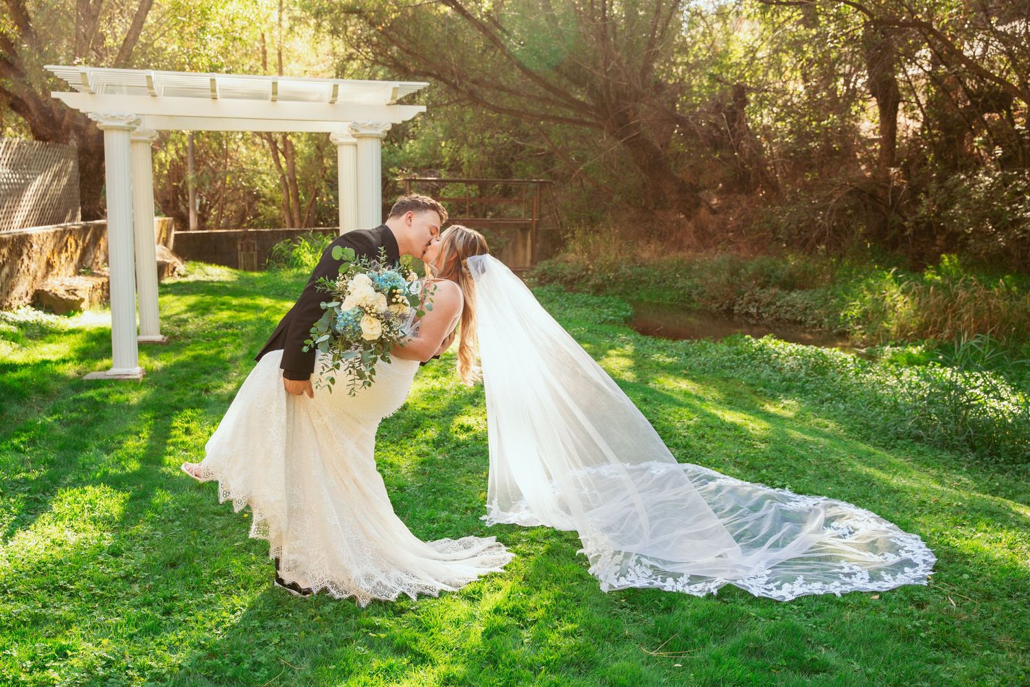 Wedding couple poses in a garden setting with white pillars and lush greenery.
