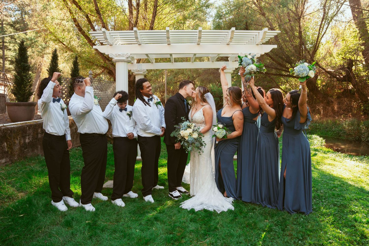 Full wedding party poses under white pergola with bridesmaids in blue and groomsmen in white shirts.