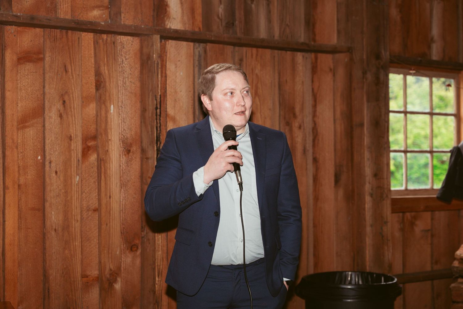 Guest in formal navy suit gestures while standing in wooden barn interior.