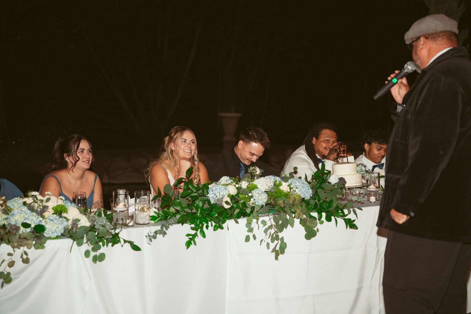 White floral and greenery decorations adorn tables at an outdoor wedding reception.