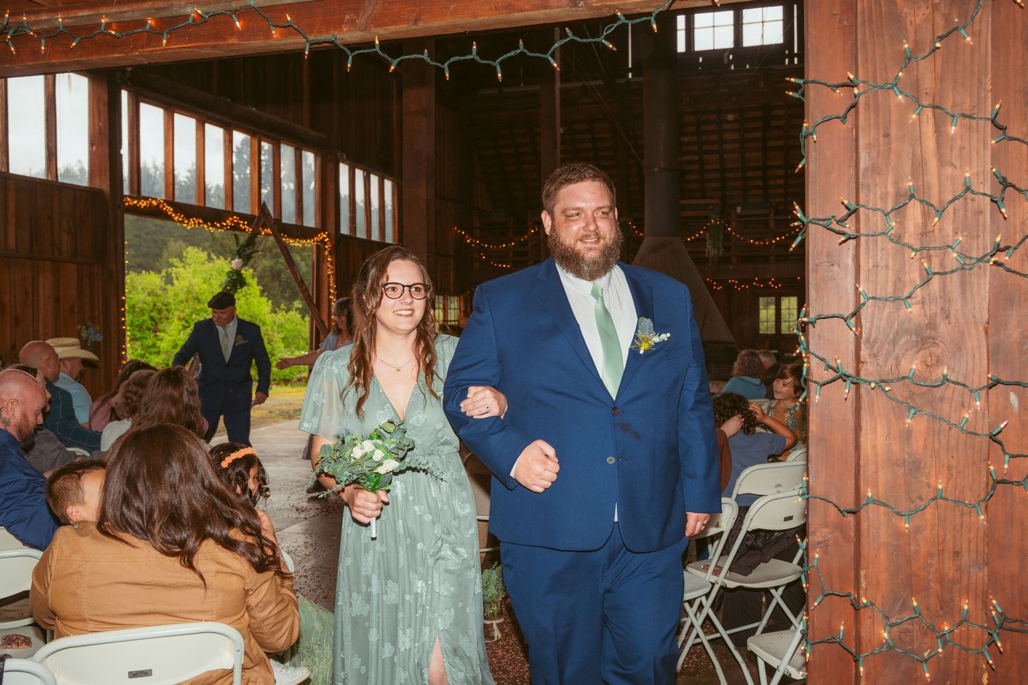 Wedding procession walking down the aisle in a rustic barn venue with string lights and wooden beams.