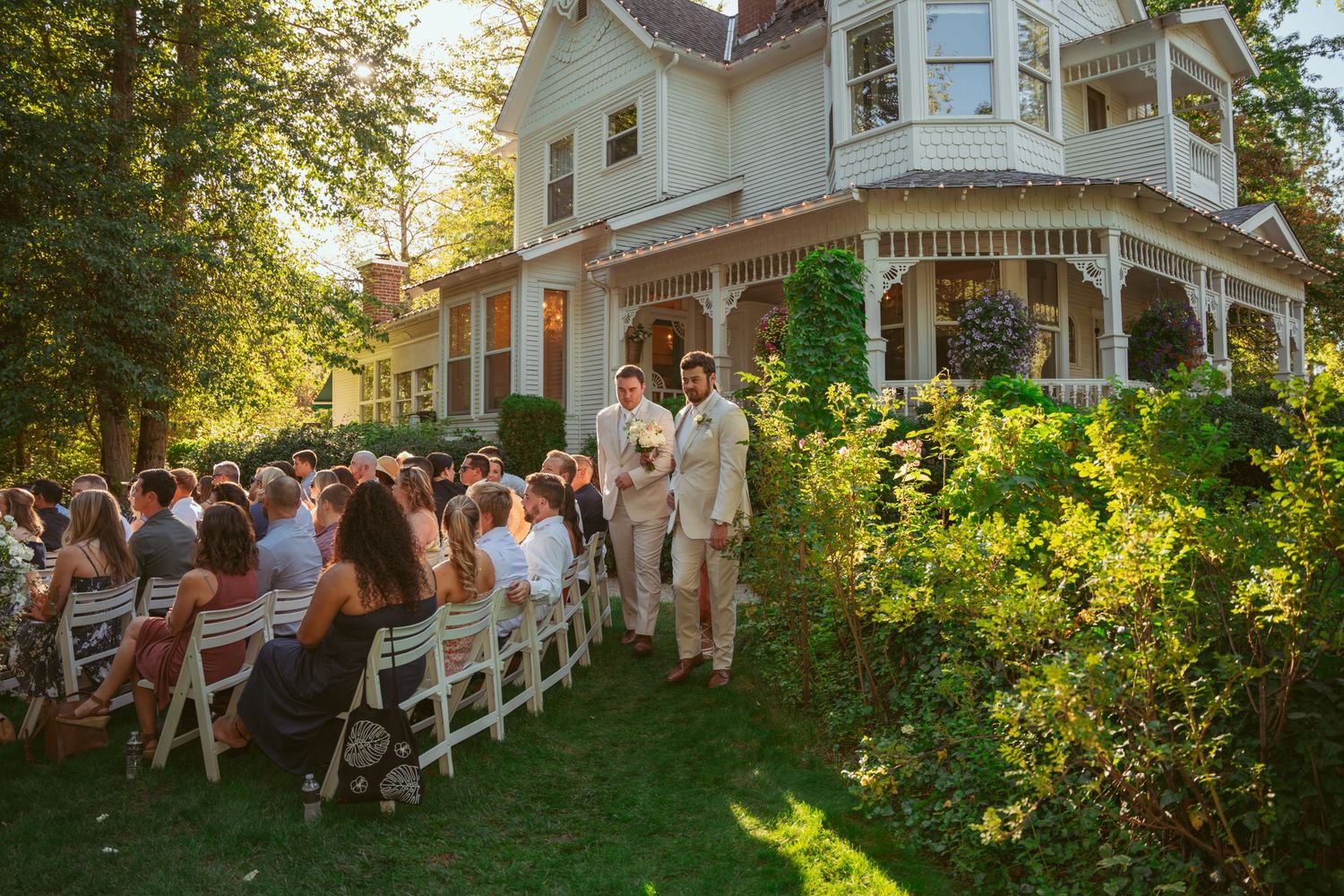Guests watch a wedding ceremony from white chairs set up on a Victorian mansion's lawn.