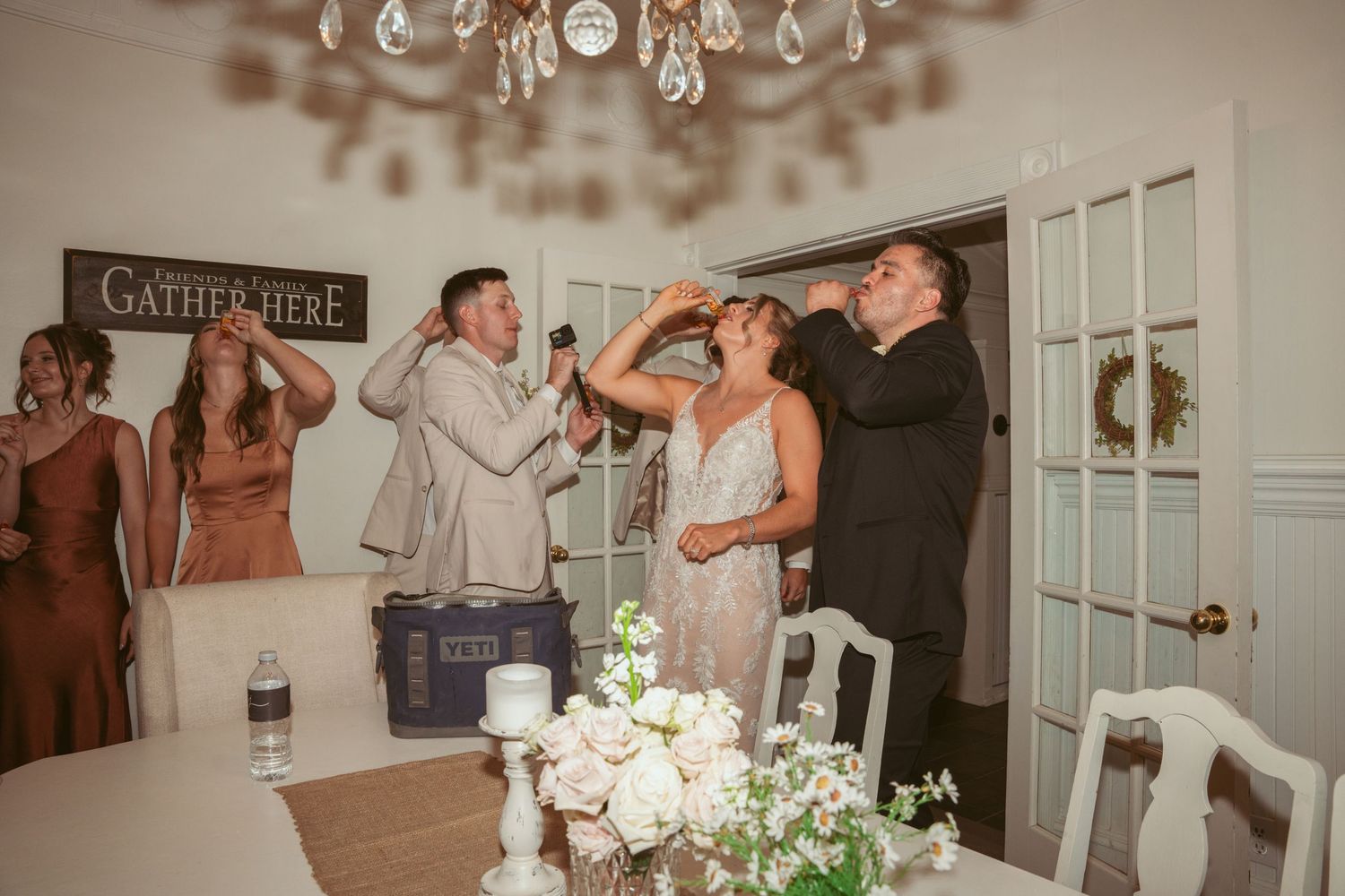 Wedding reception celebration inside an elegant white room with chandelier and rustic gather sign on wall.