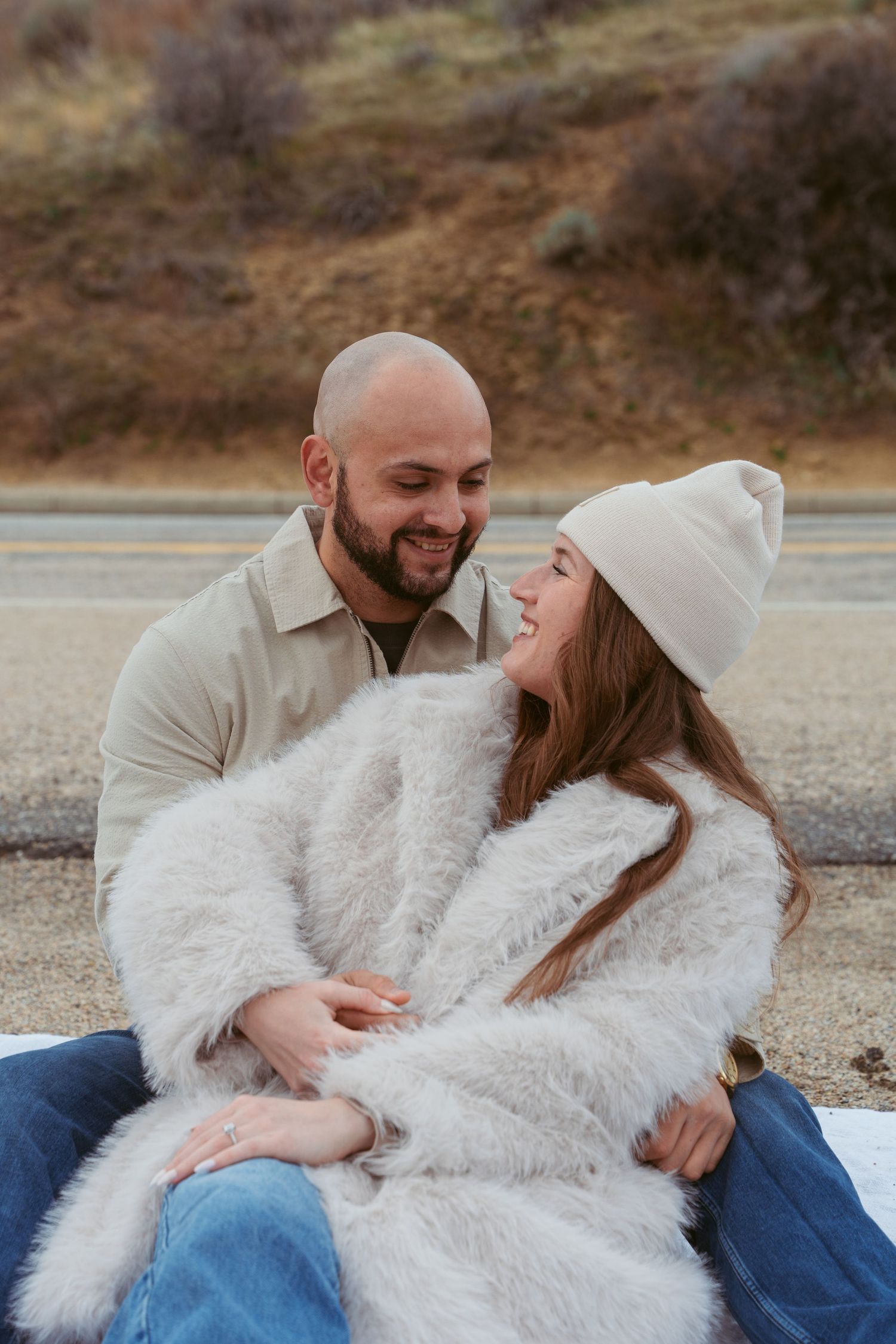 A couple shares an intimate moment together while sitting on the ground in casual winter attire.