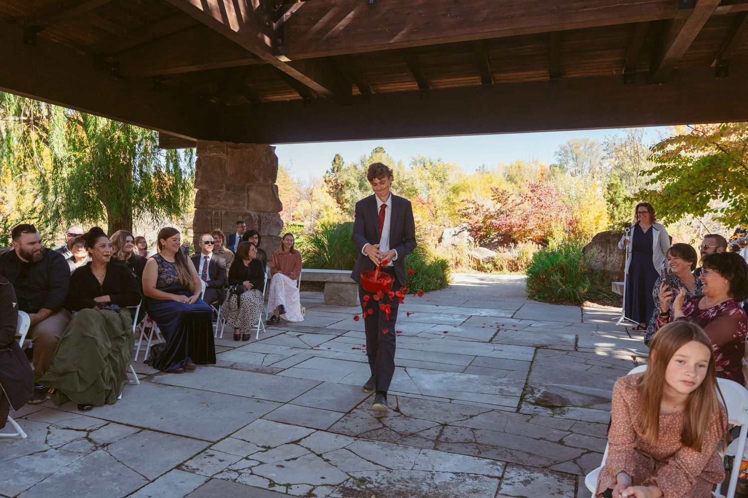 Wedding ceremony attendees gather under a covered stone patio surrounded by fall foliage.