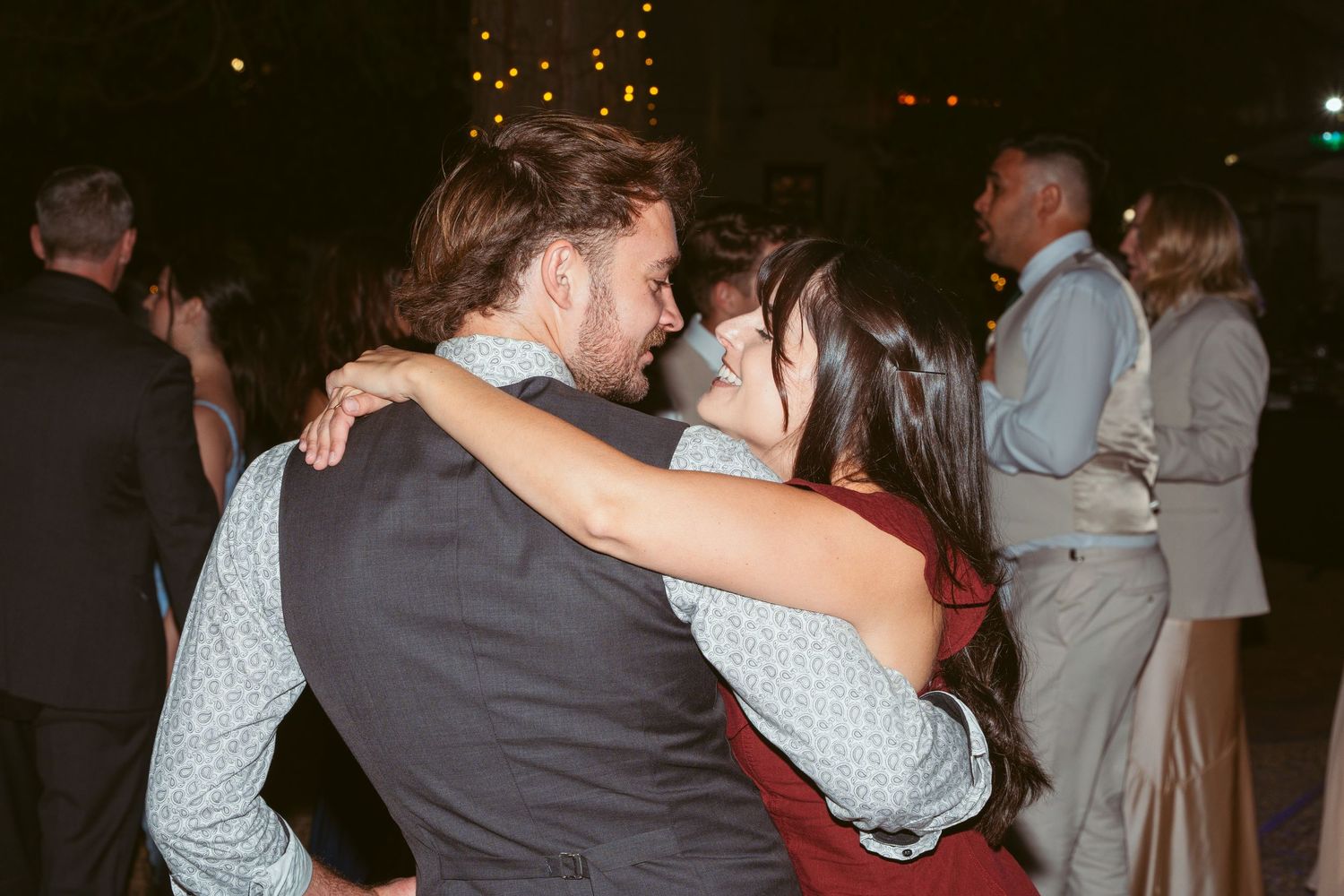 Couples dancing together at a dimly lit outdoor wedding reception with string lights overhead.