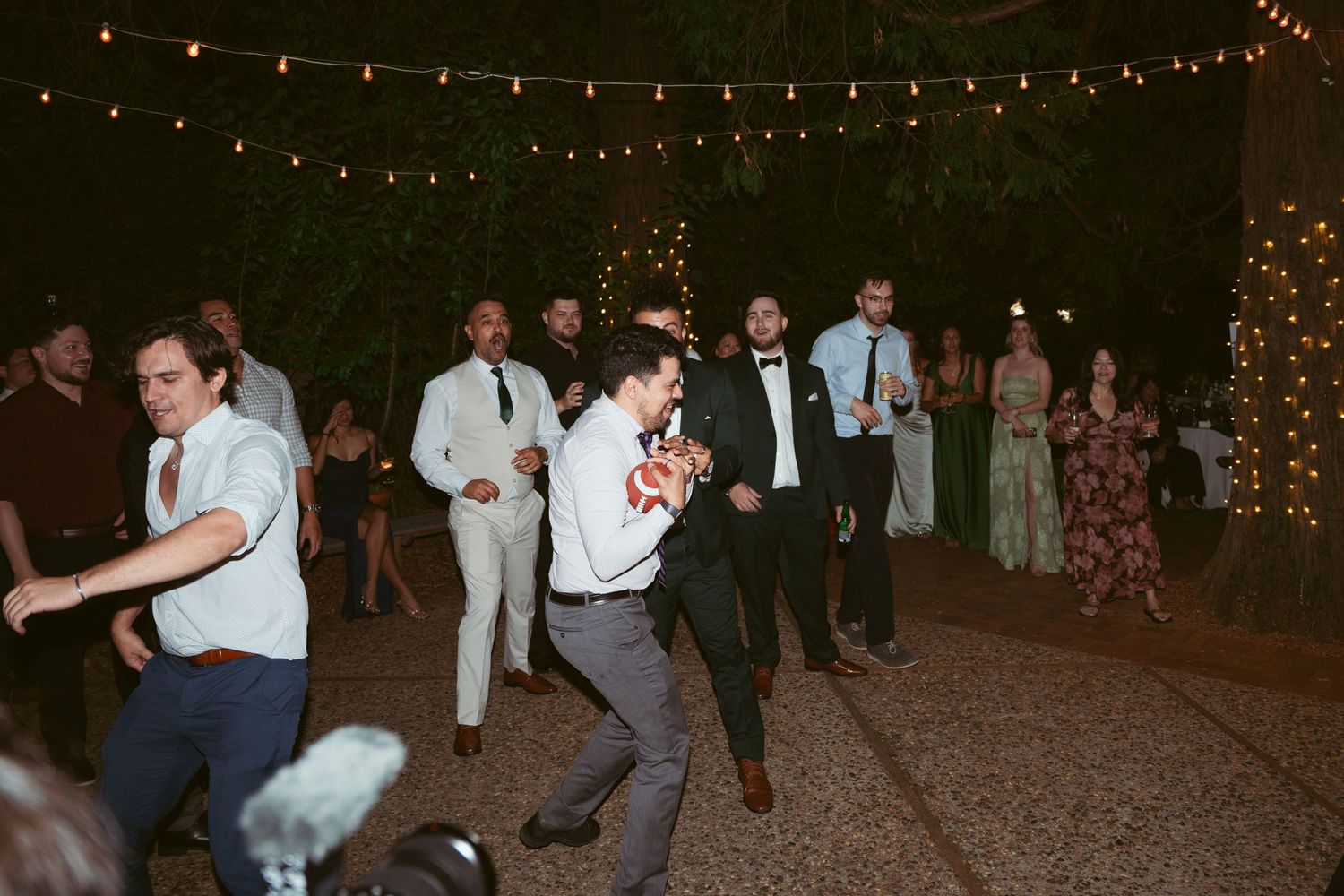 Wedding guests dancing under string lights at an outdoor evening reception.