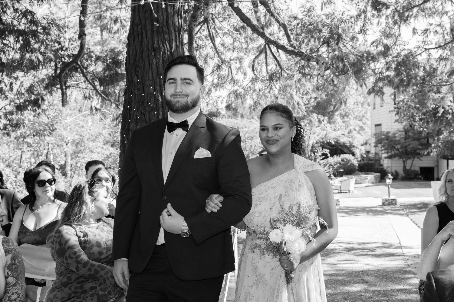 A black and white photo of a wedding ceremony in a park setting with guests looking on during a sunny day.