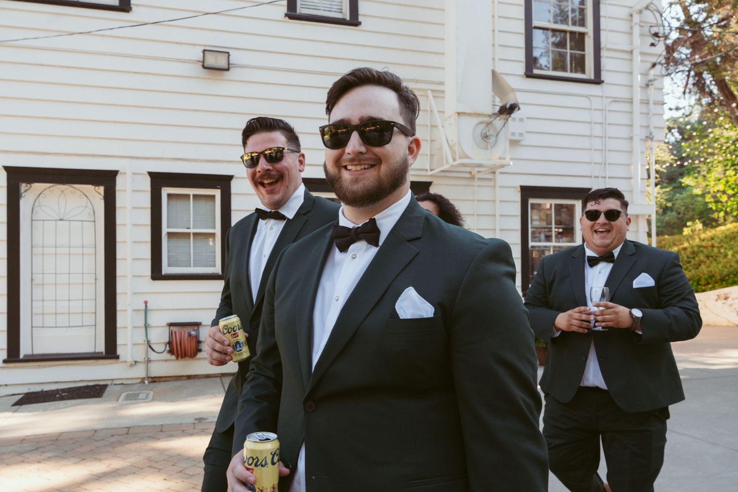 Groomsmen in formal attire and sunglasses pose outside a white building.