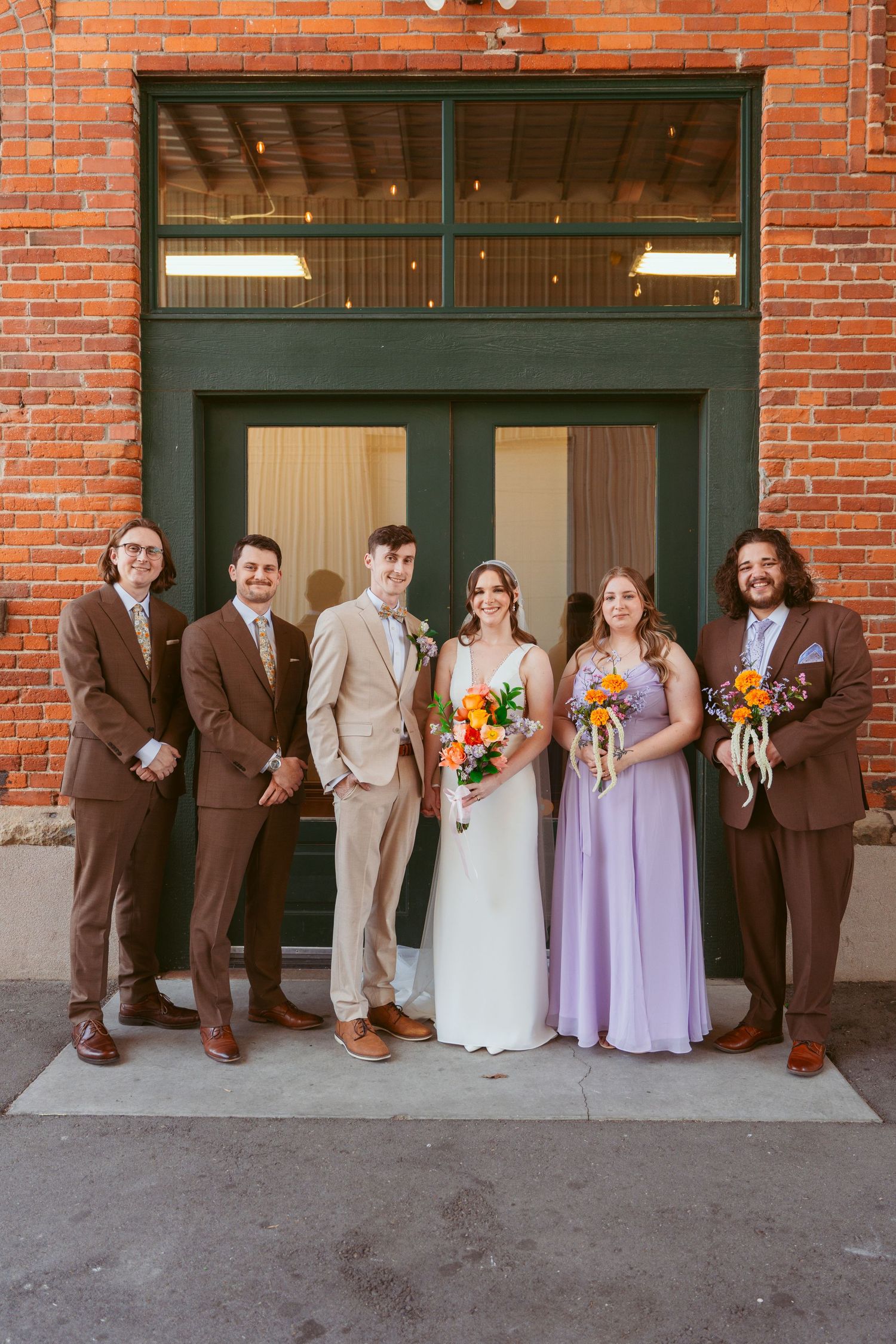 Wedding party lined up against brick building exterior in purple and brown attire.
