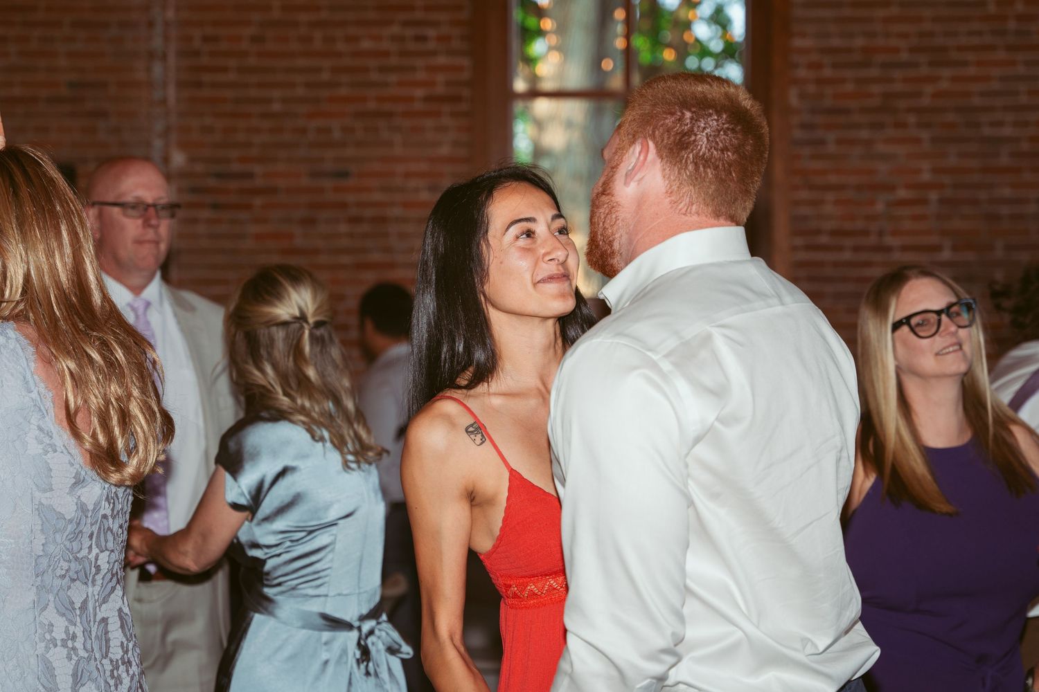 Couples dance together at an elegant wedding reception in a brick-walled venue with natural lighting.