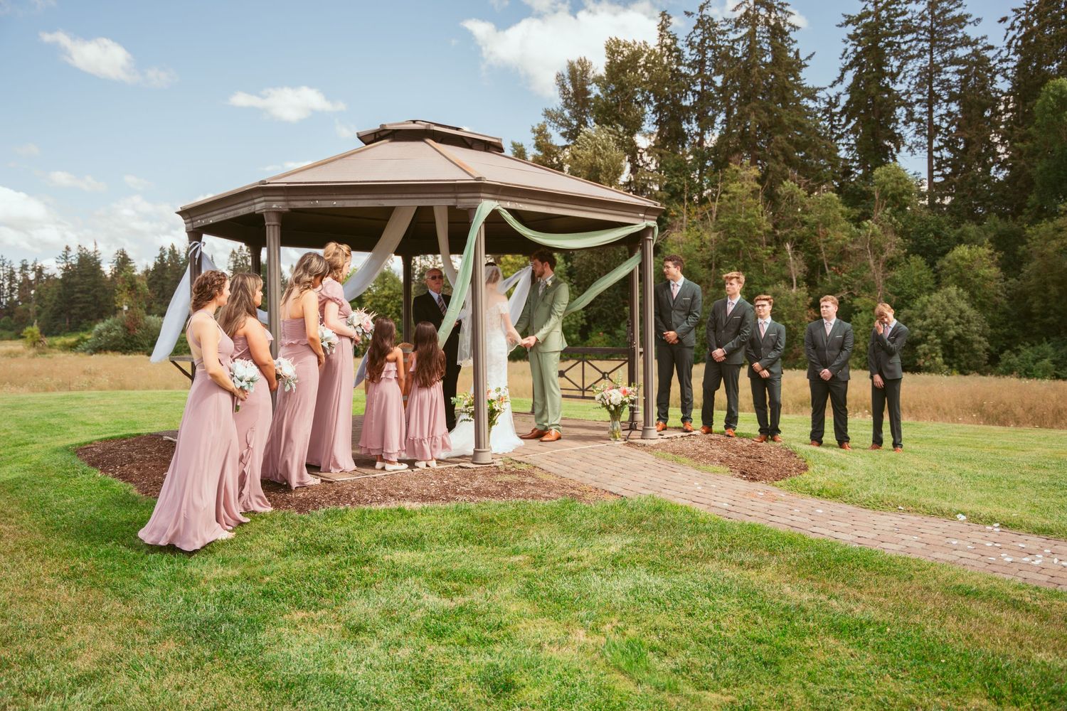Wedding party in pink and green attire poses under wooden gazebo in outdoor rustic setting.