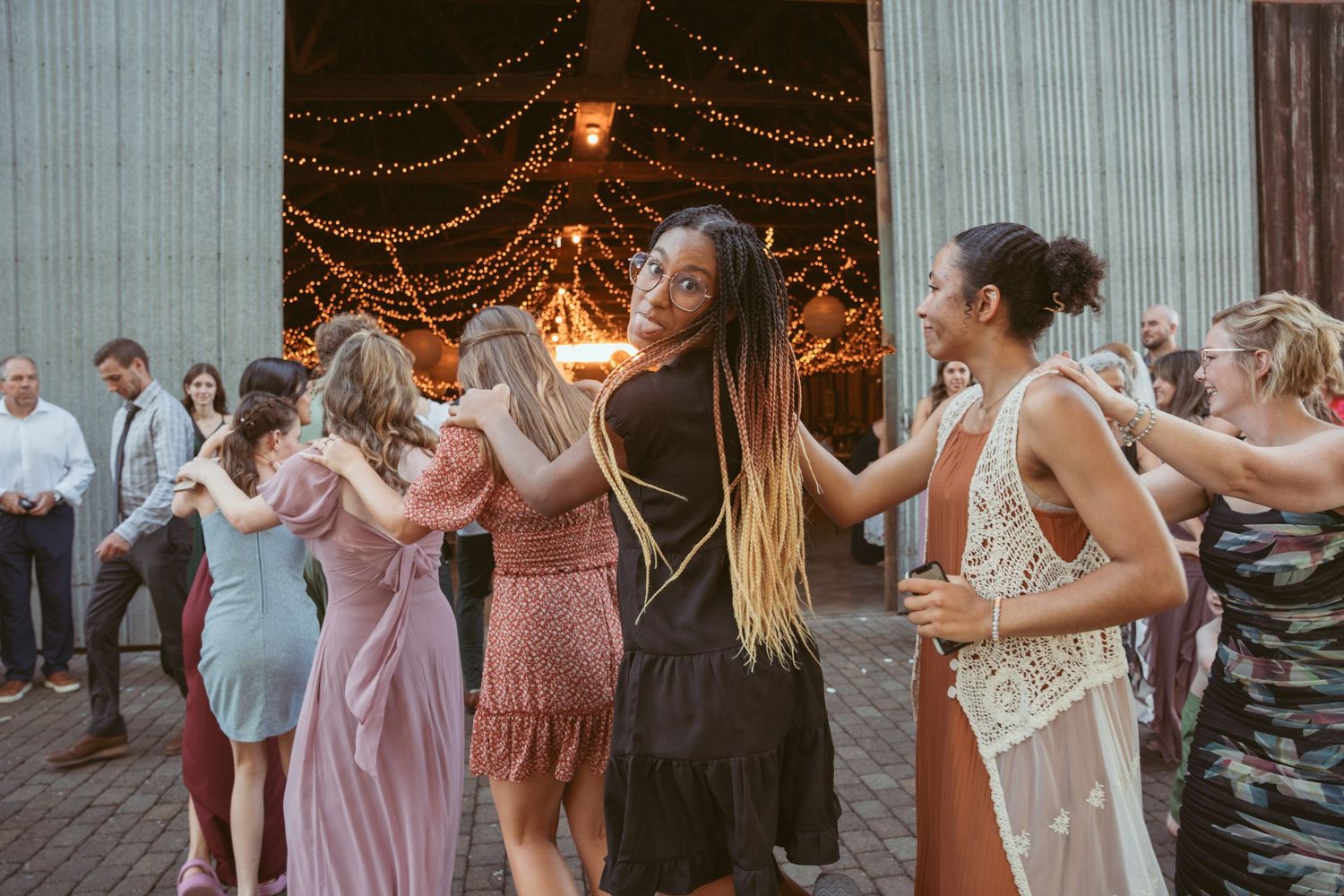 Wedding guests participate in group dance formation under romantic string lights.