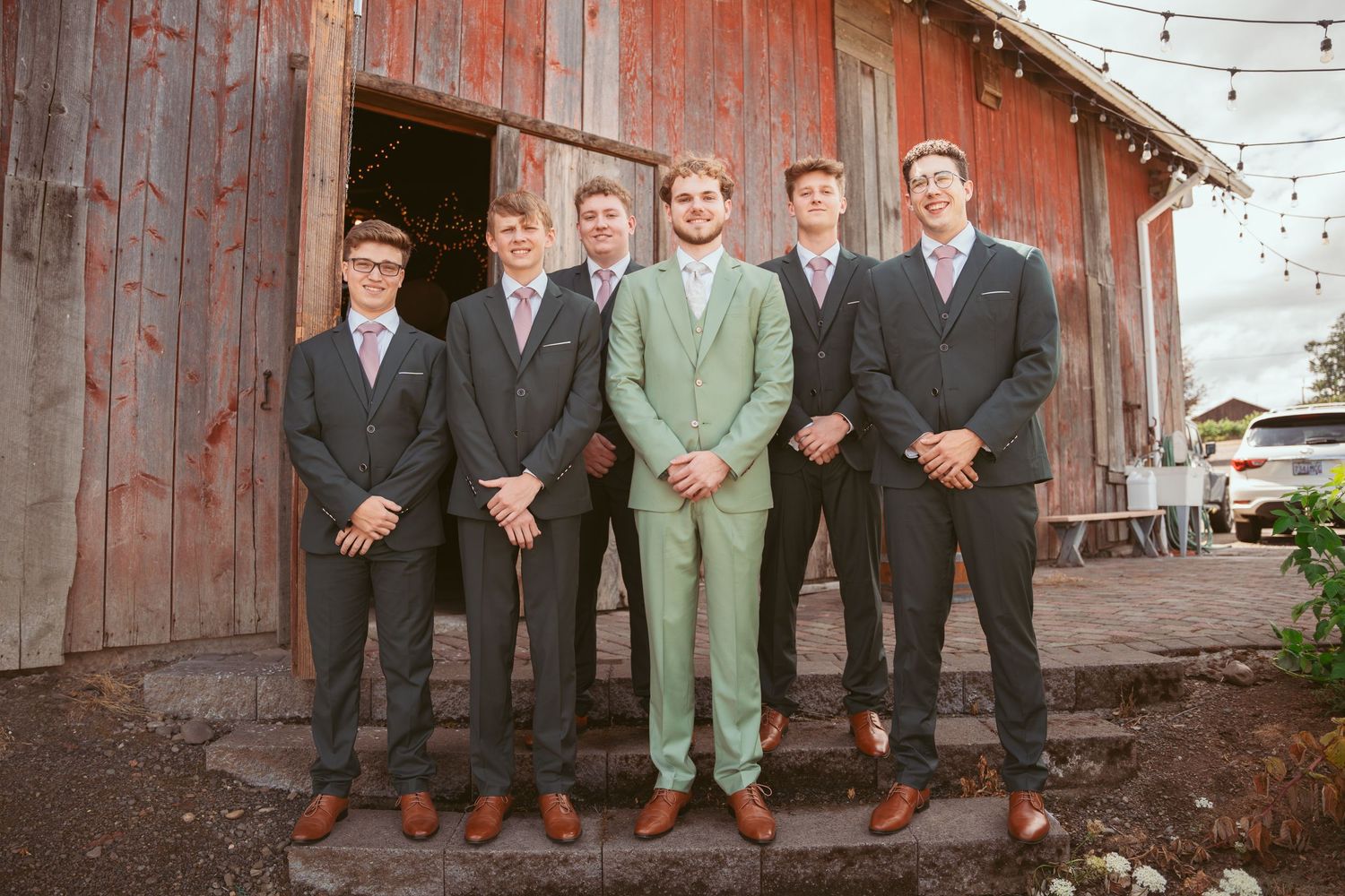 Wedding party poses in front of rustic red barn with string lights.