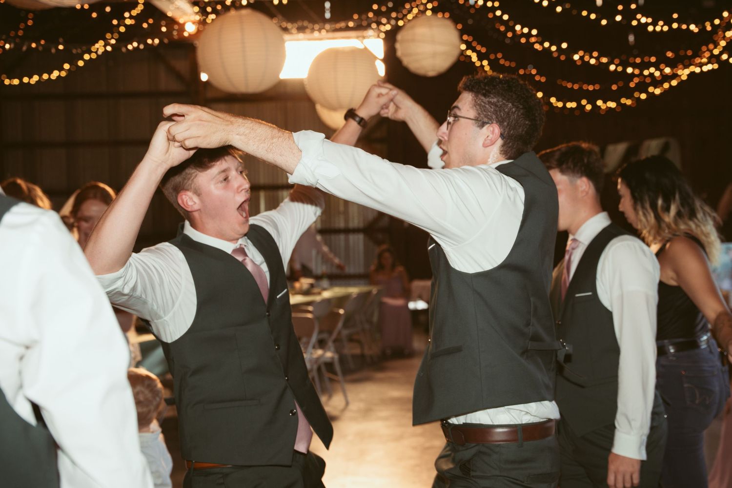 Groomsmen in vests celebrate with high-fives on the dance floor under string lights.