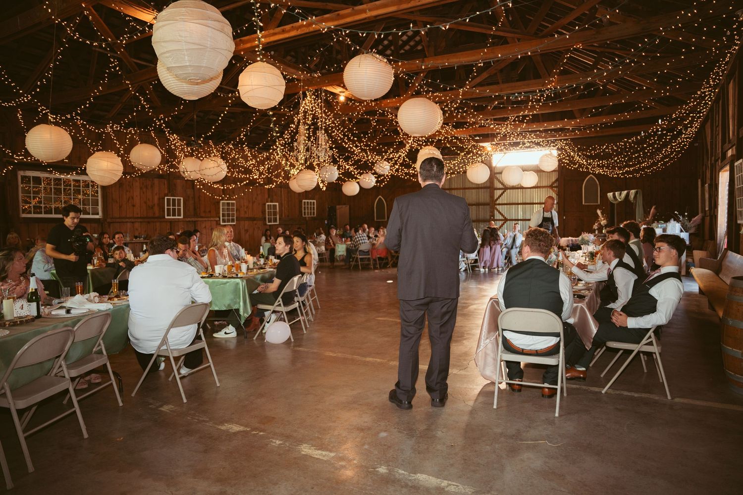 Wedding reception venue decorated with string lights and paper lanterns in rustic barn setting.