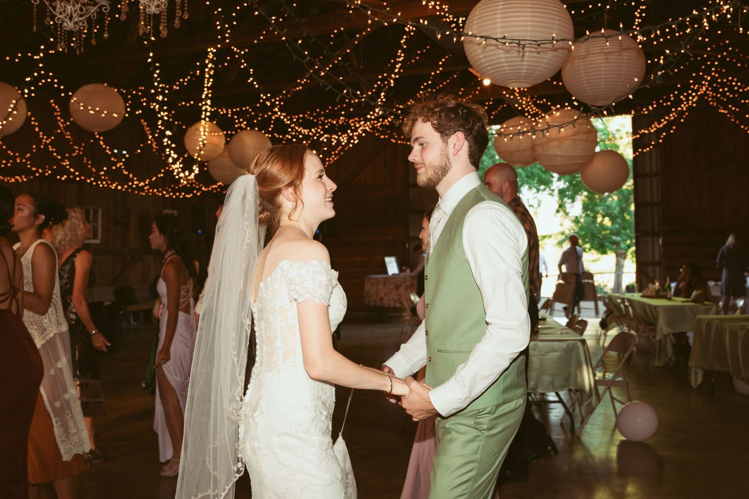 Wedding party dancing and celebrating under romantic string light decorations in a rustic venue.