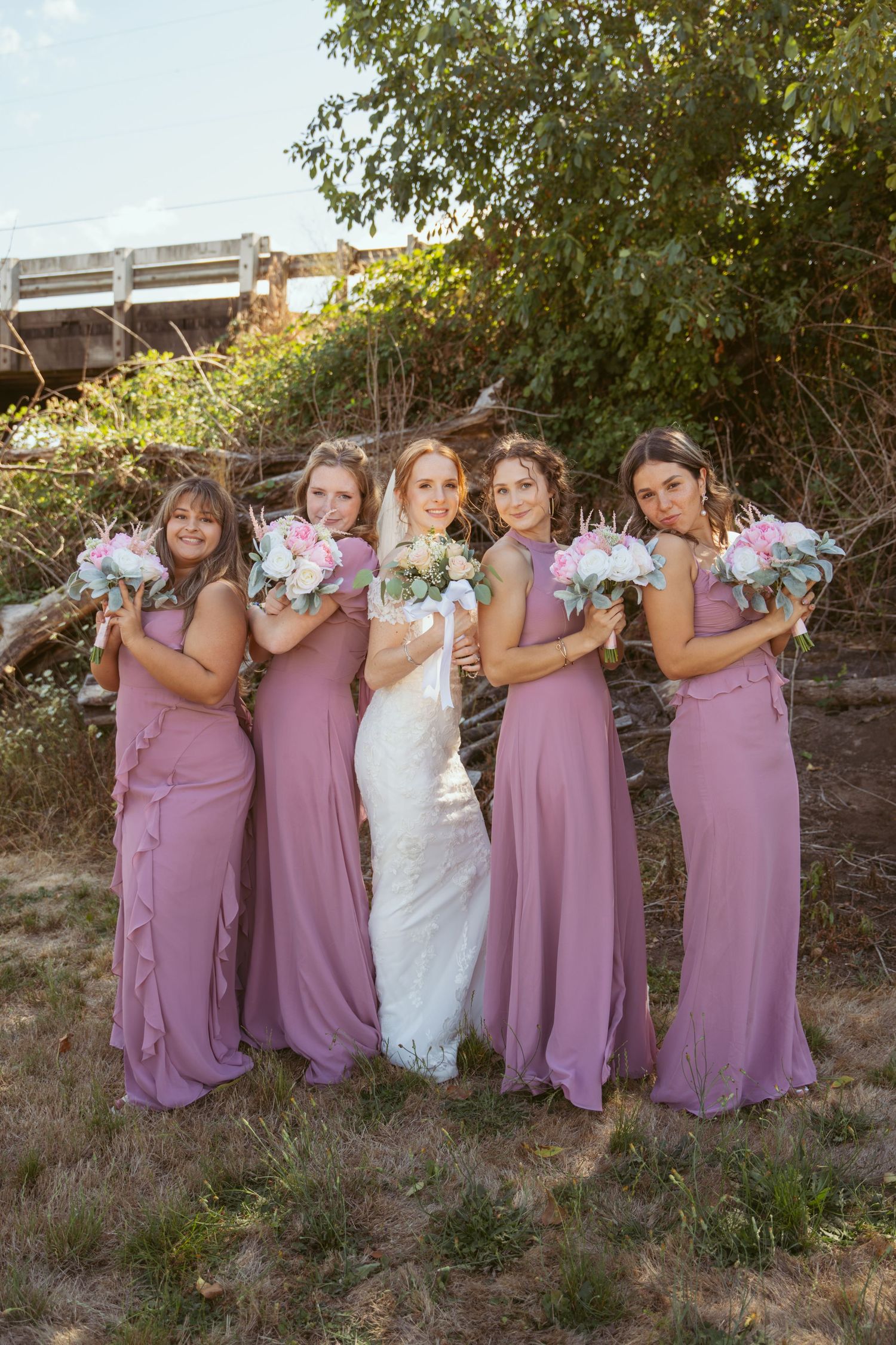 Wedding party in mauve dresses and white gown pose playfully with bouquets outdoors.