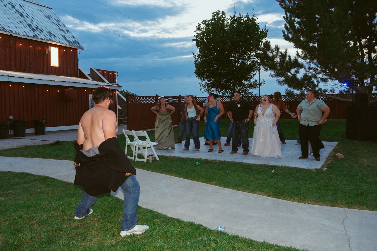 Outdoor wedding celebration with guests dancing near a red barn under blue skies.