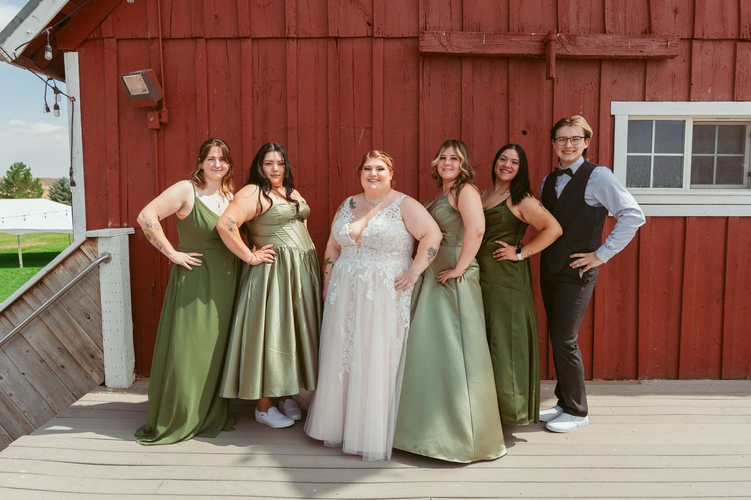 Bridesmaids in sage green dresses pose with bride in white gown against red barn exterior.