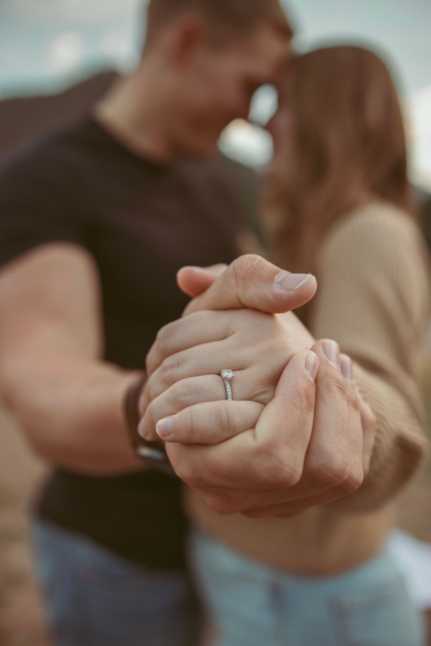 Close-up of clasped hands showing an engagement ring against a blurred romantic background.