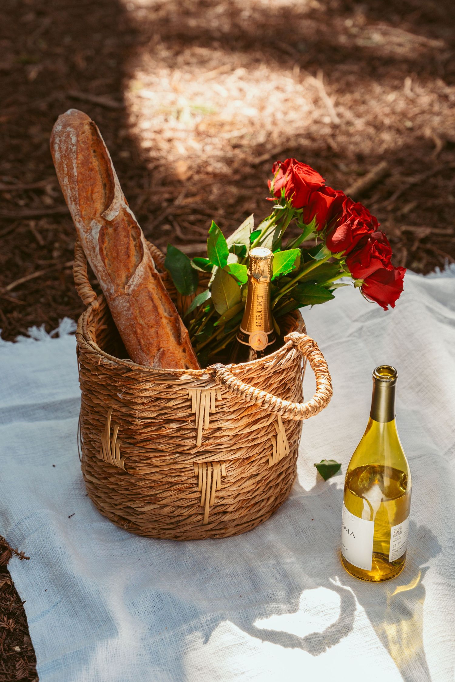 Wicker picnic basket with wine bottles and red roses arranged on white blanket in dappled sunlight.