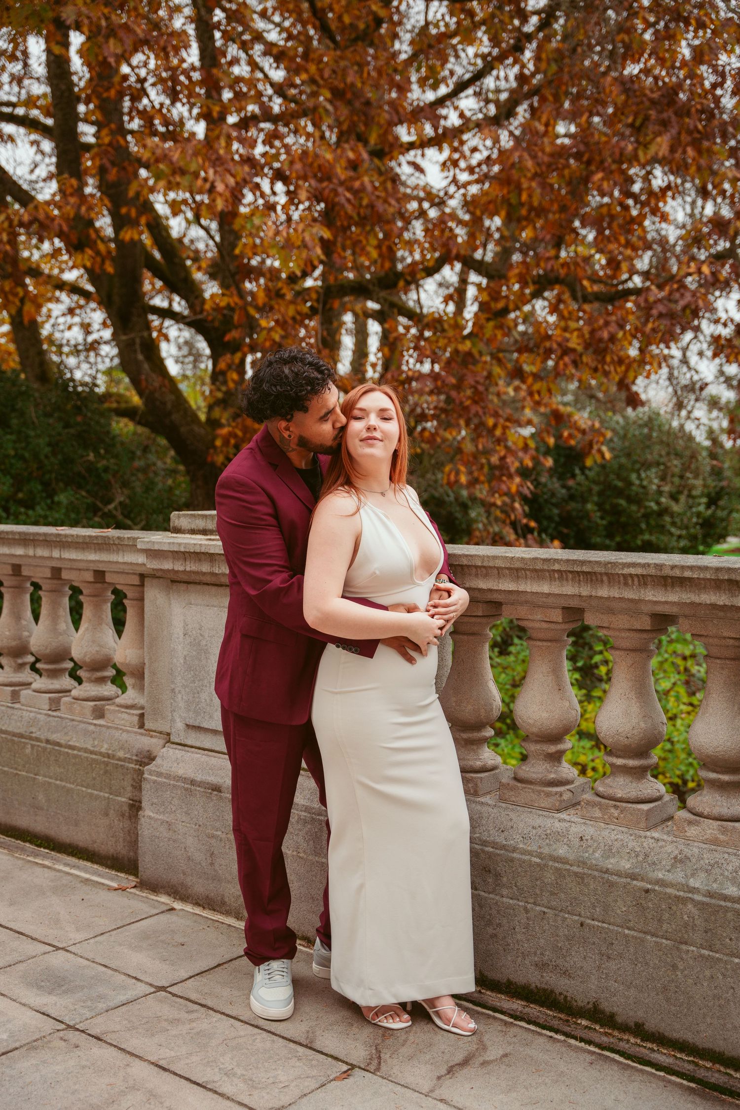 A couple embraces on a stone balcony surrounded by fall foliage and ornate balusters.