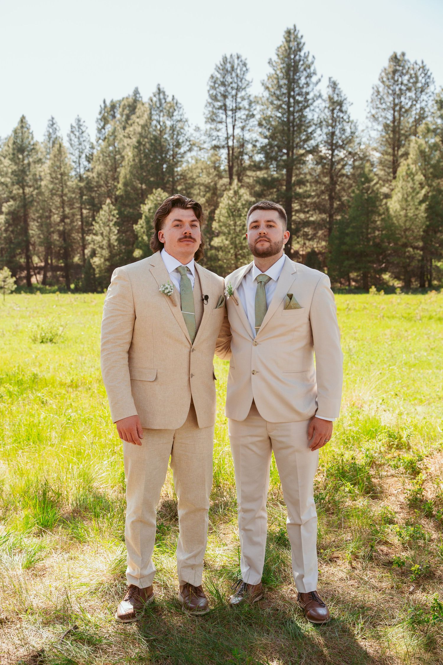 Two people in matching tan suits stand together in a sunlit field near pine trees.