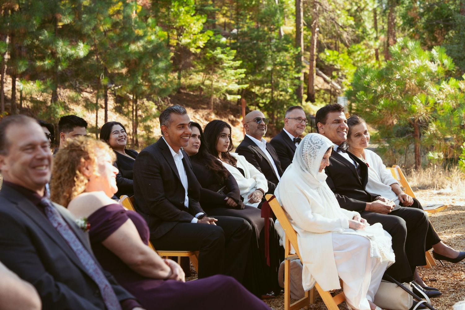 Wedding attendees gather in a forest setting with natural lighting filtering through the trees.