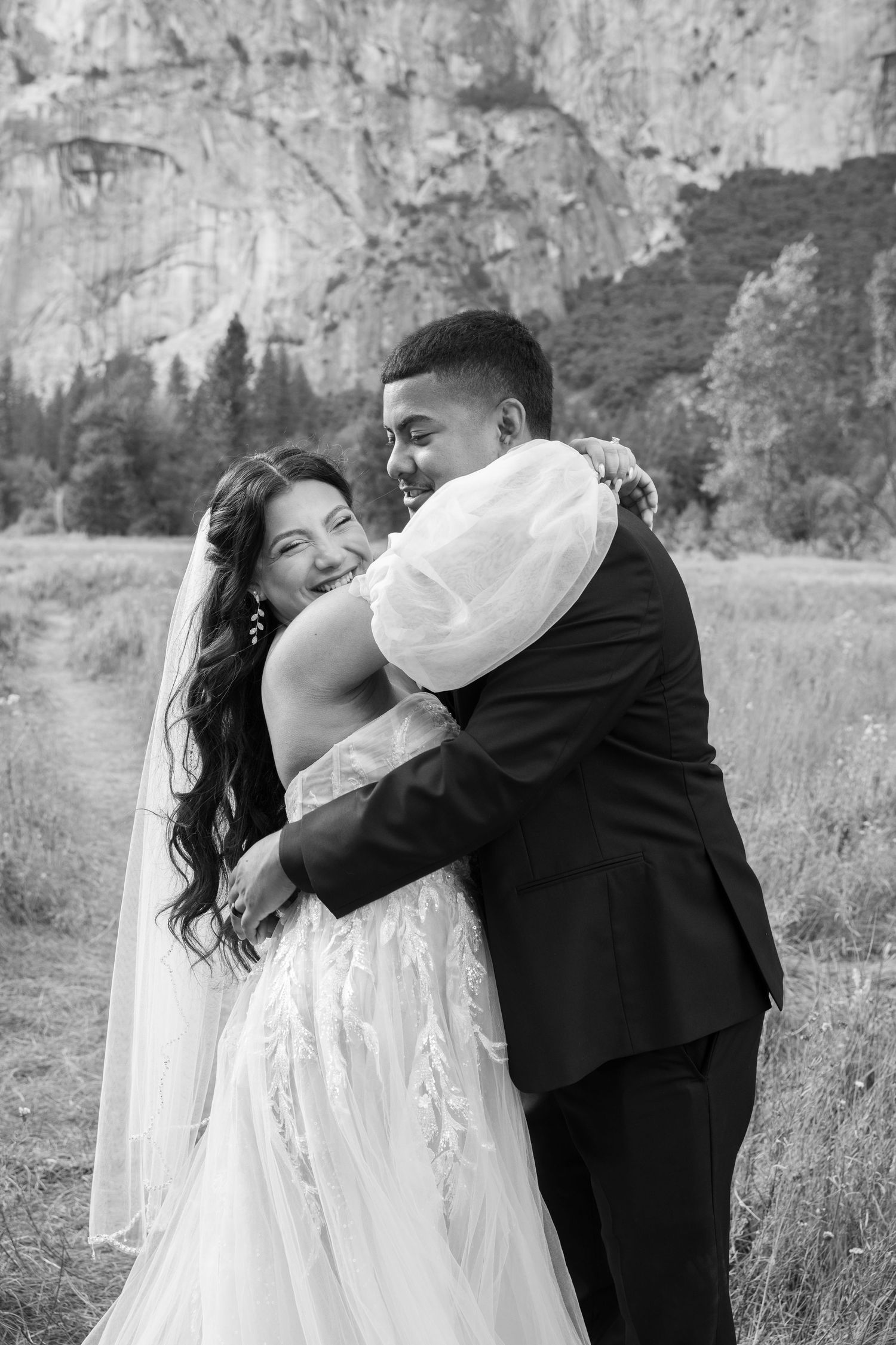 A romantic wedding couple shares a kiss in front of dramatic mountain cliffs in black and white.