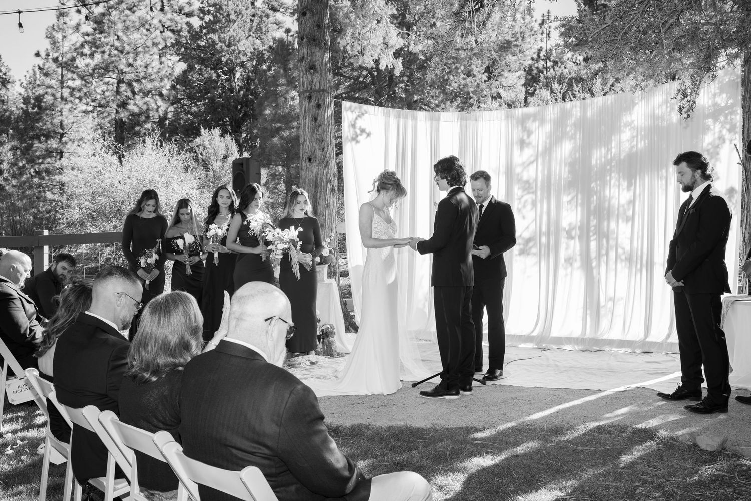 Black and white photo of outdoor wedding ceremony with guests seated on white chairs.
