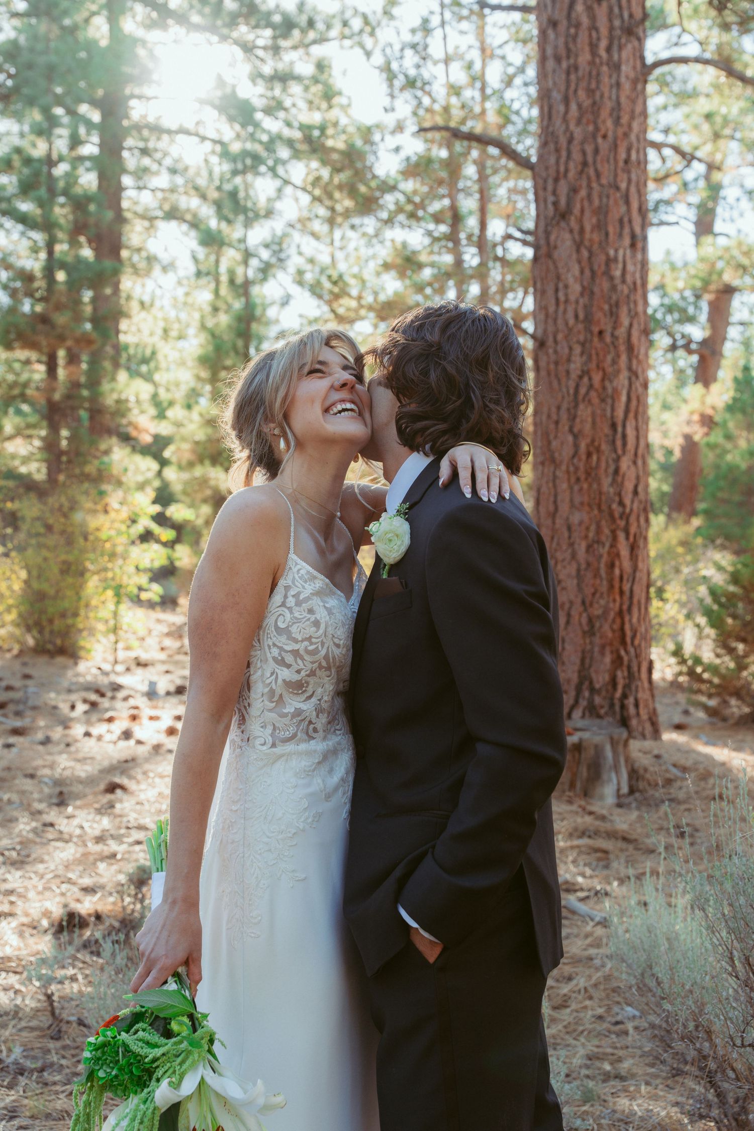 A couple embraces during their wedding photoshoot in a sunlit pine forest.