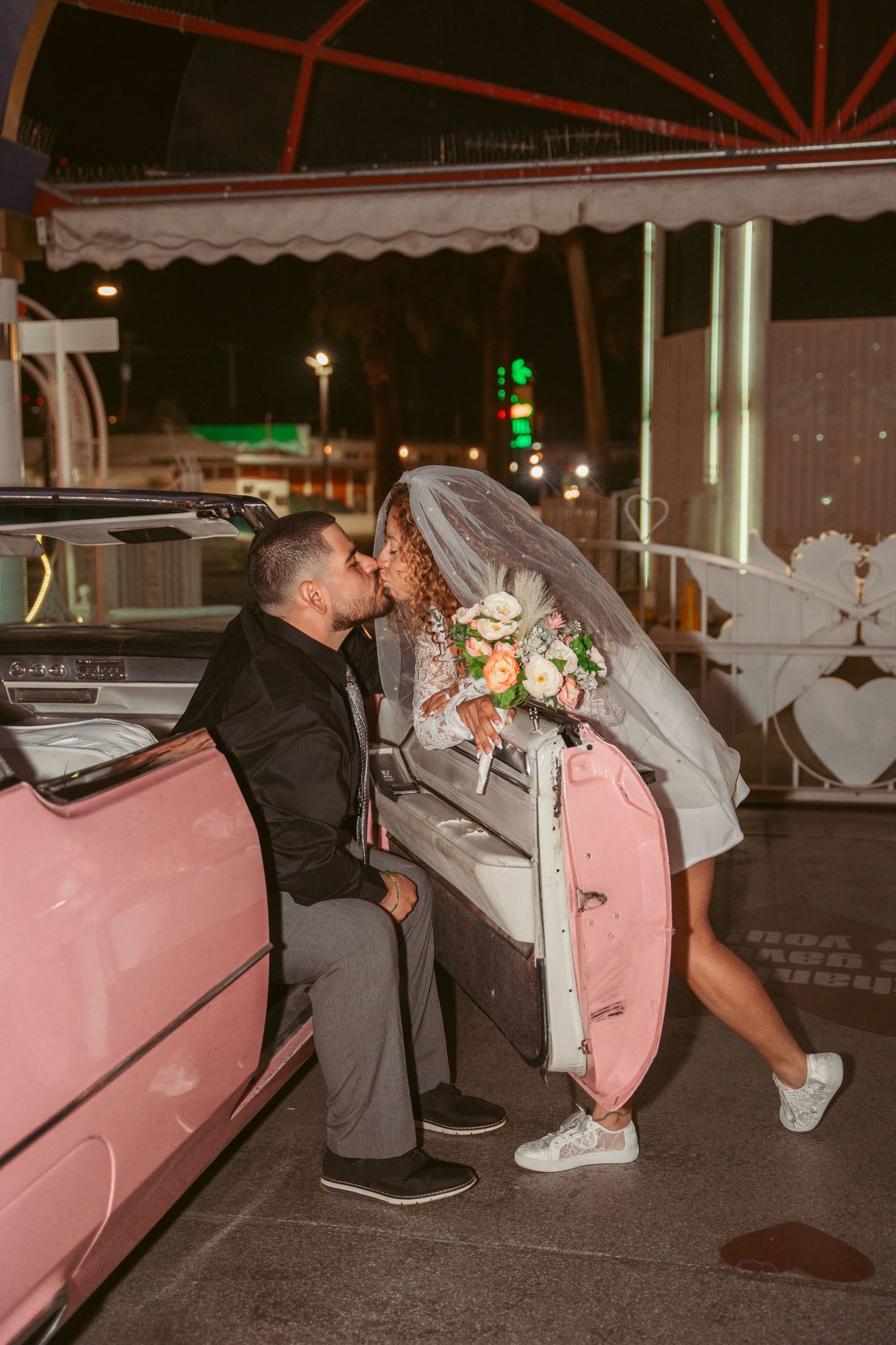 Couple sharing a romantic kiss in front of a pink vintage car at night.