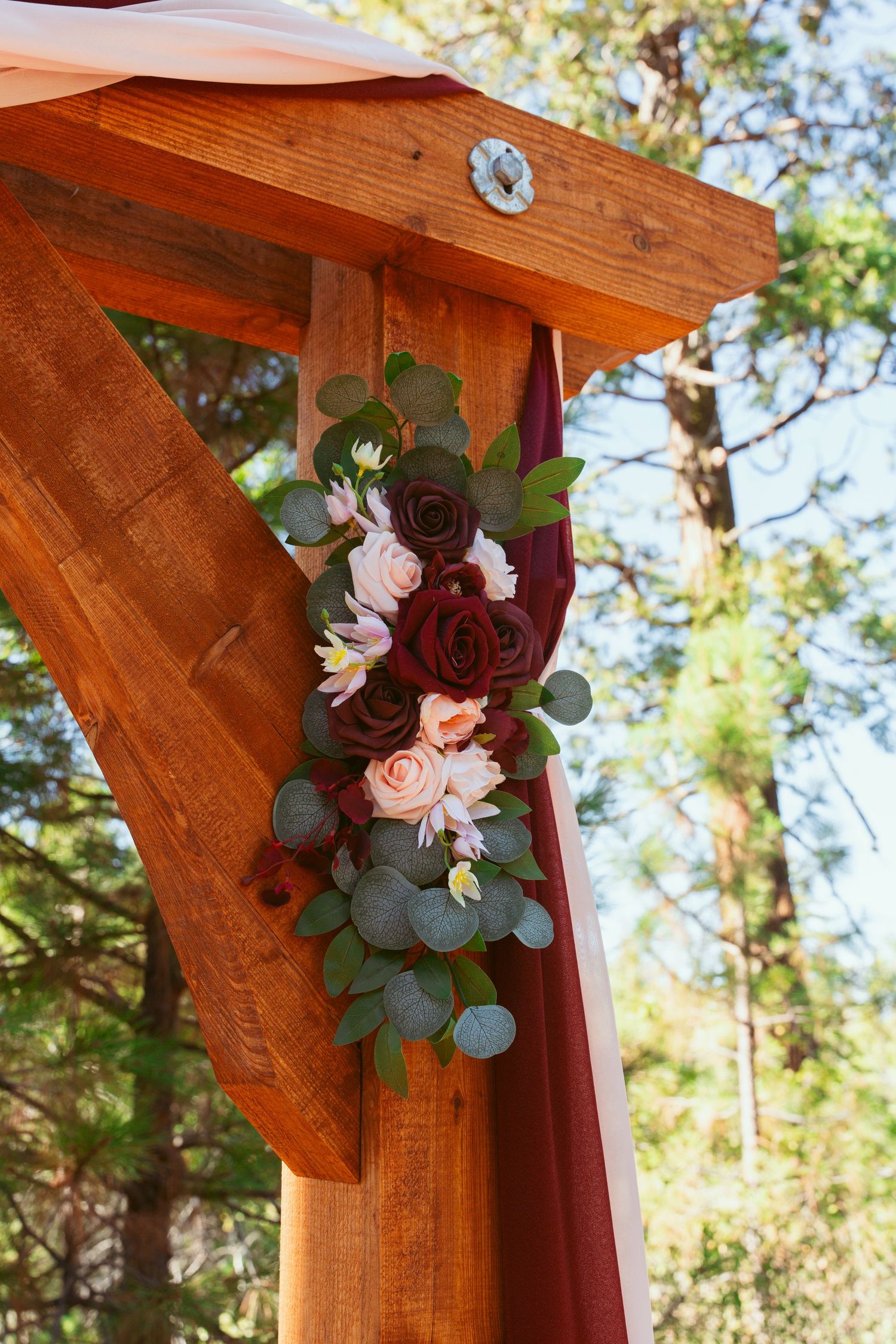 Wooden arch detail decorated with burgundy roses and eucalyptus in outdoor setting.