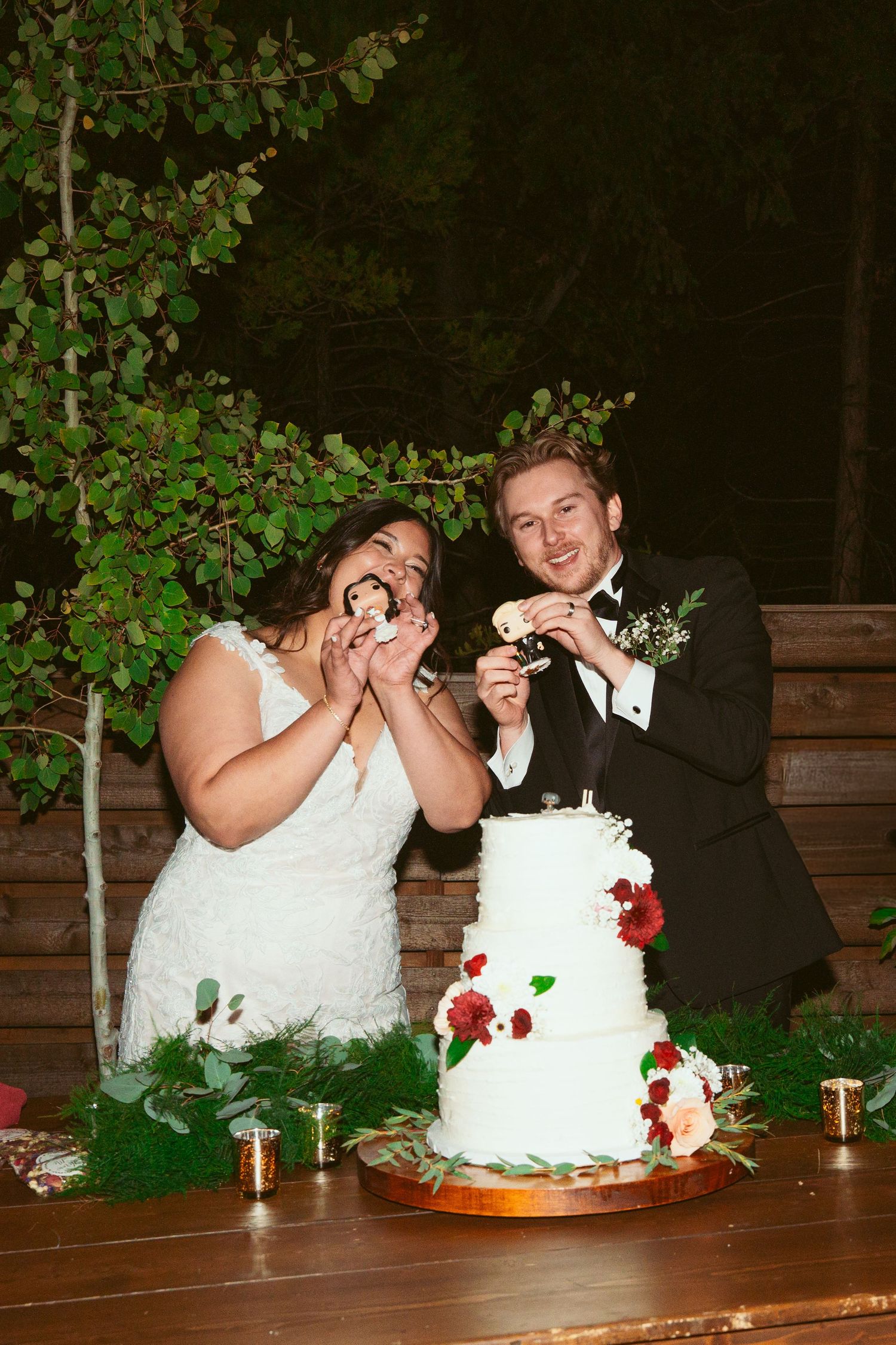 Couple shares wedding cake during reception with decorative greenery in background.