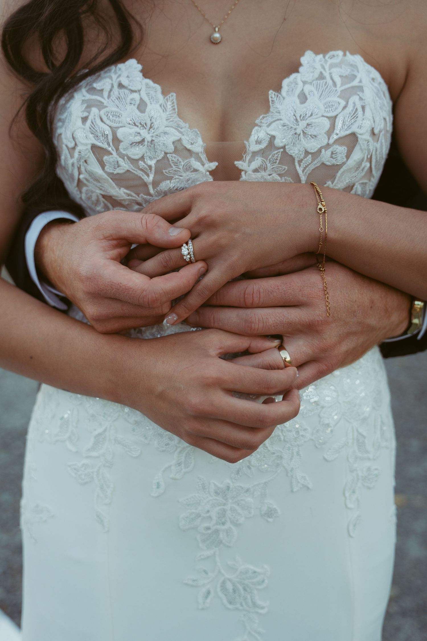 Detail shot of intricate lace wedding dress bodice with jewelry and hands showing rings.