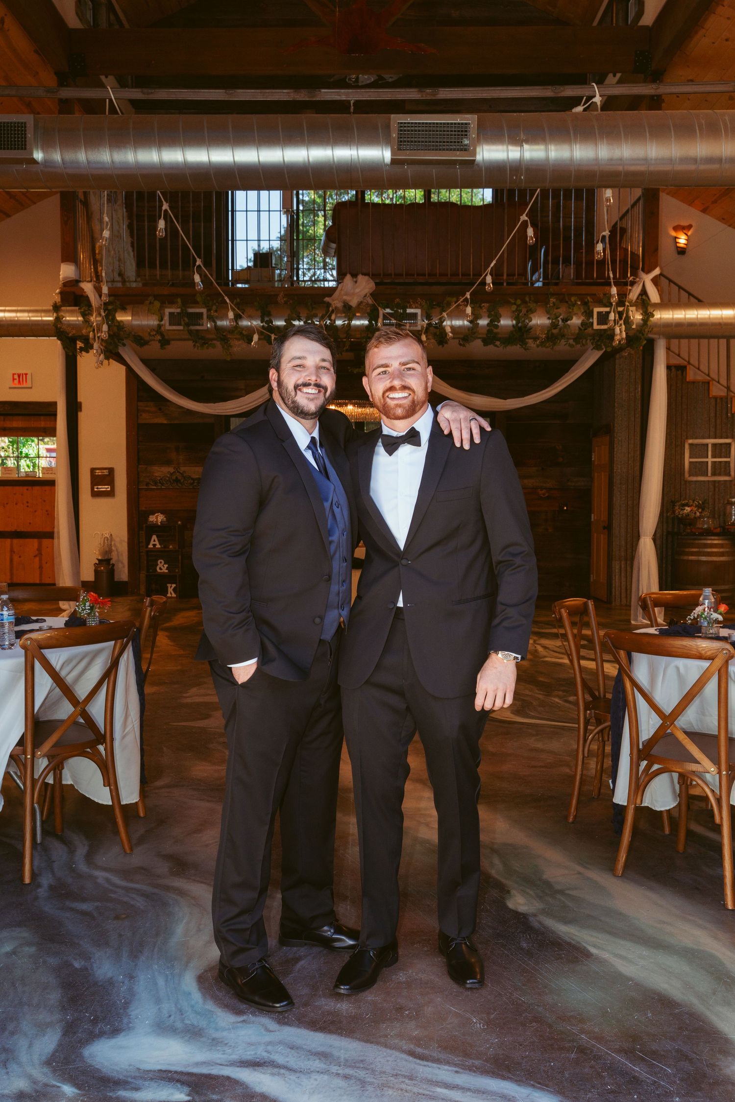 Two men in formal attire smiling at a rustic indoor wedding reception venue.