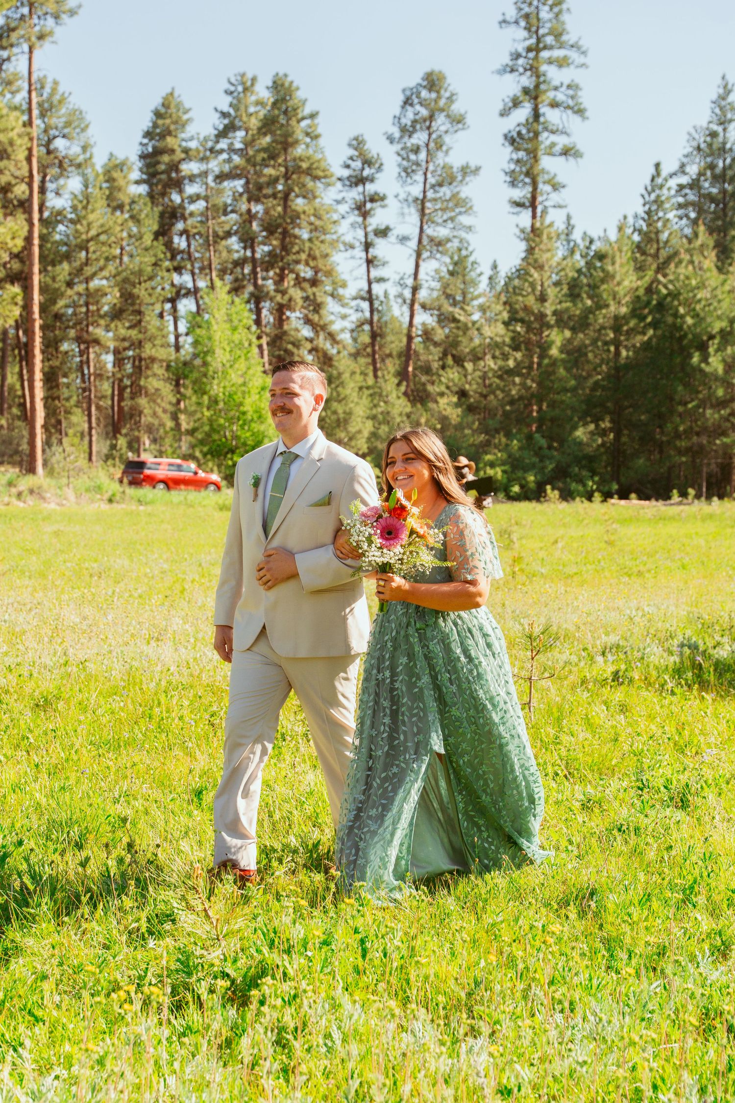 Wedding party members strolling through a sunlit meadow surrounded by tall pine trees.