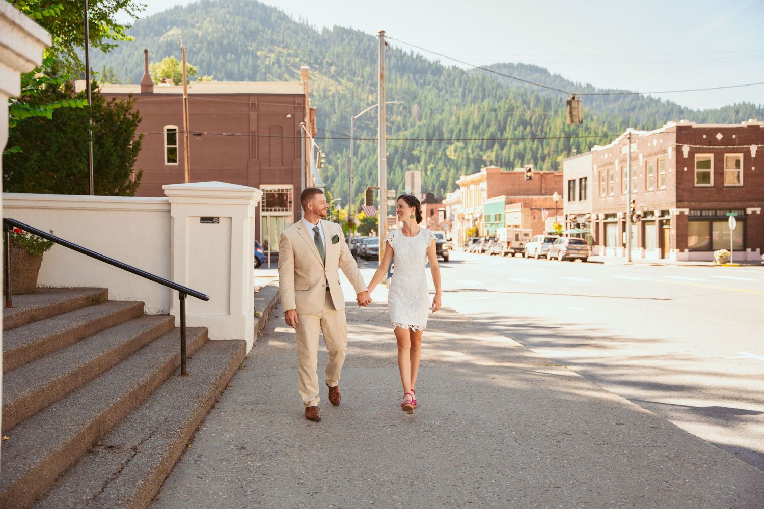 A couple walks down a sunny small-town street with mountains in the background.