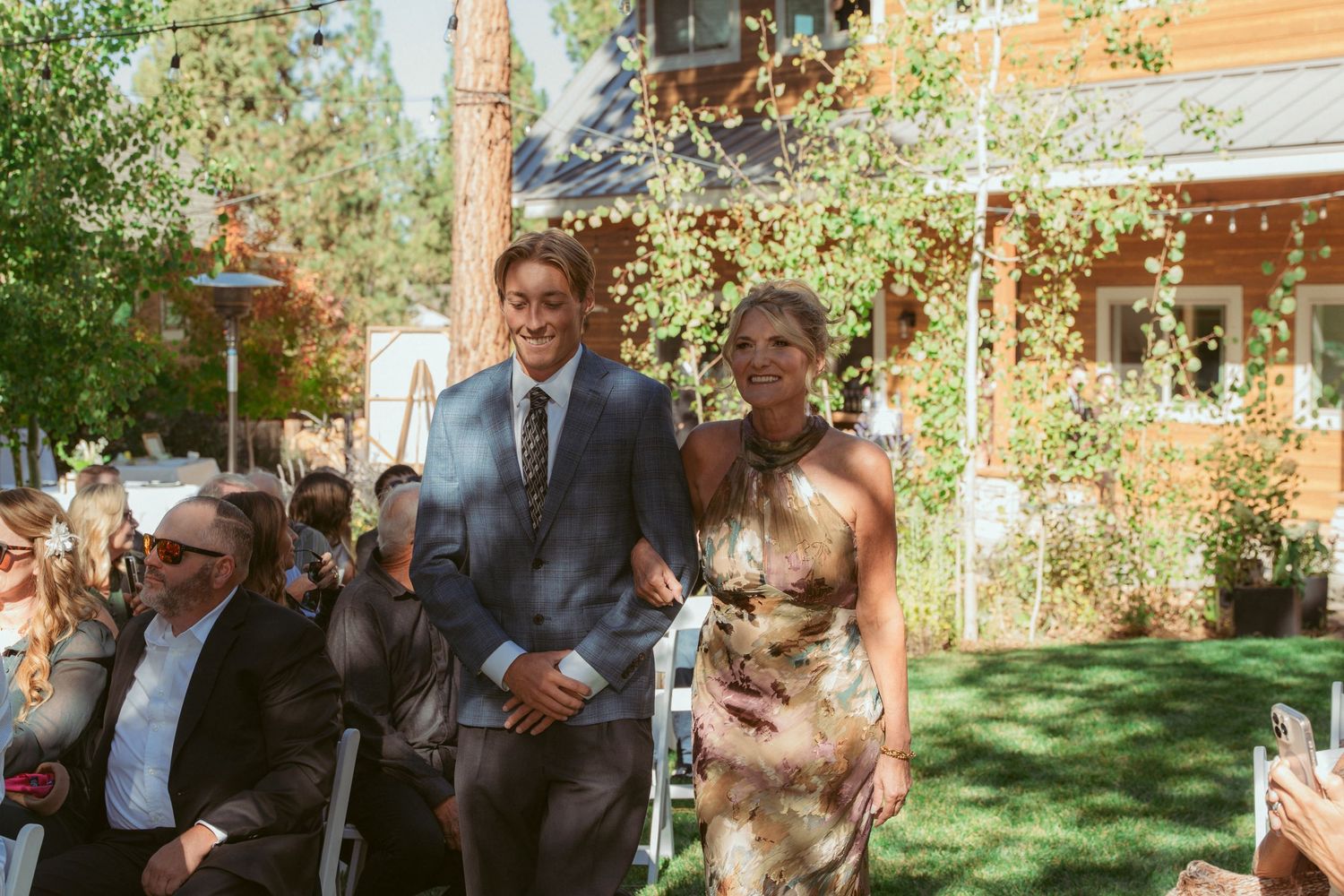 Wedding processional on a sunny lawn with rustic cabin and string lights in background.