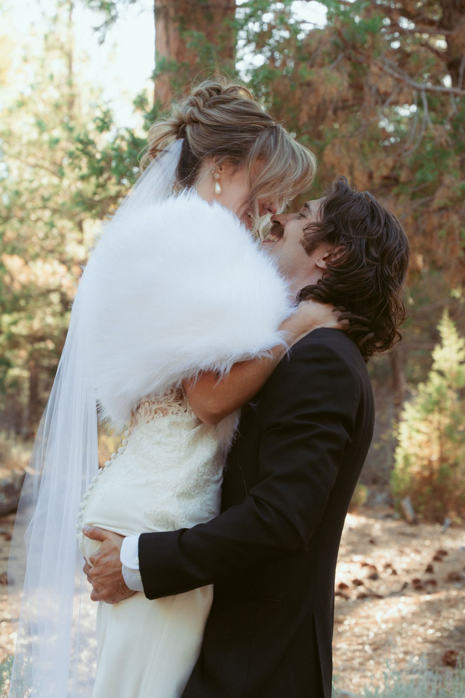Newlyweds share an intimate moment among tall pine trees in an outdoor forest setting.