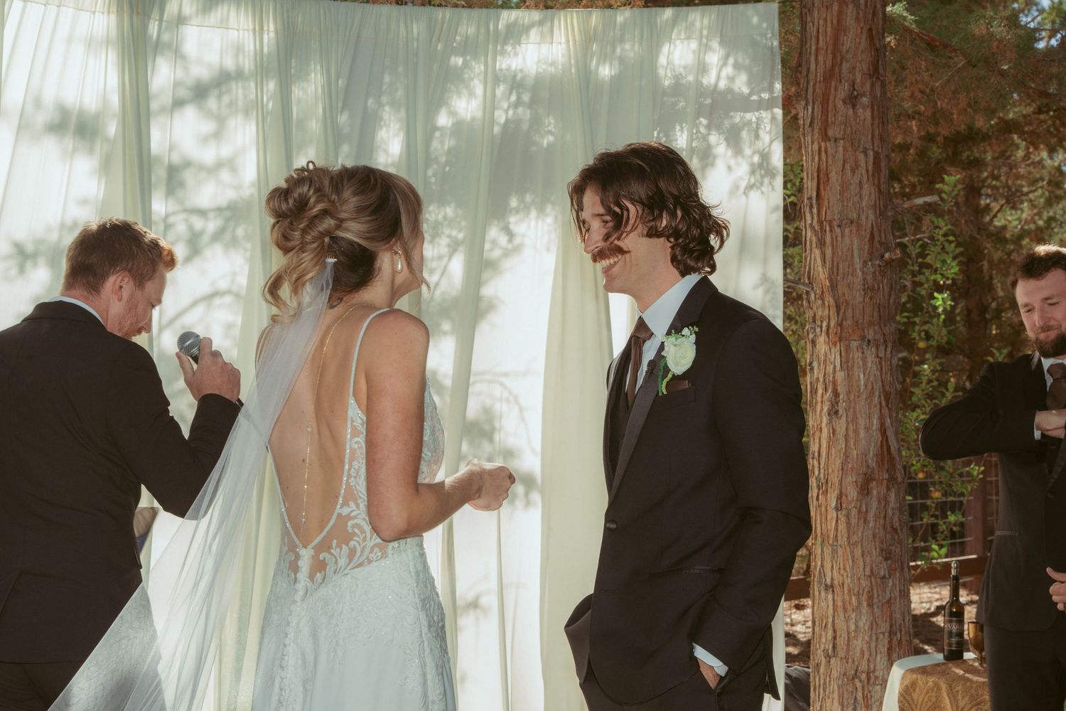 An outdoor wedding ceremony under tall trees with white fabric draping in the background.