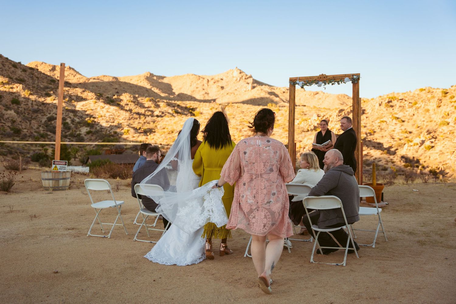 Desert wedding ceremony setup with wooden arch and white chairs against mountainous backdrop.