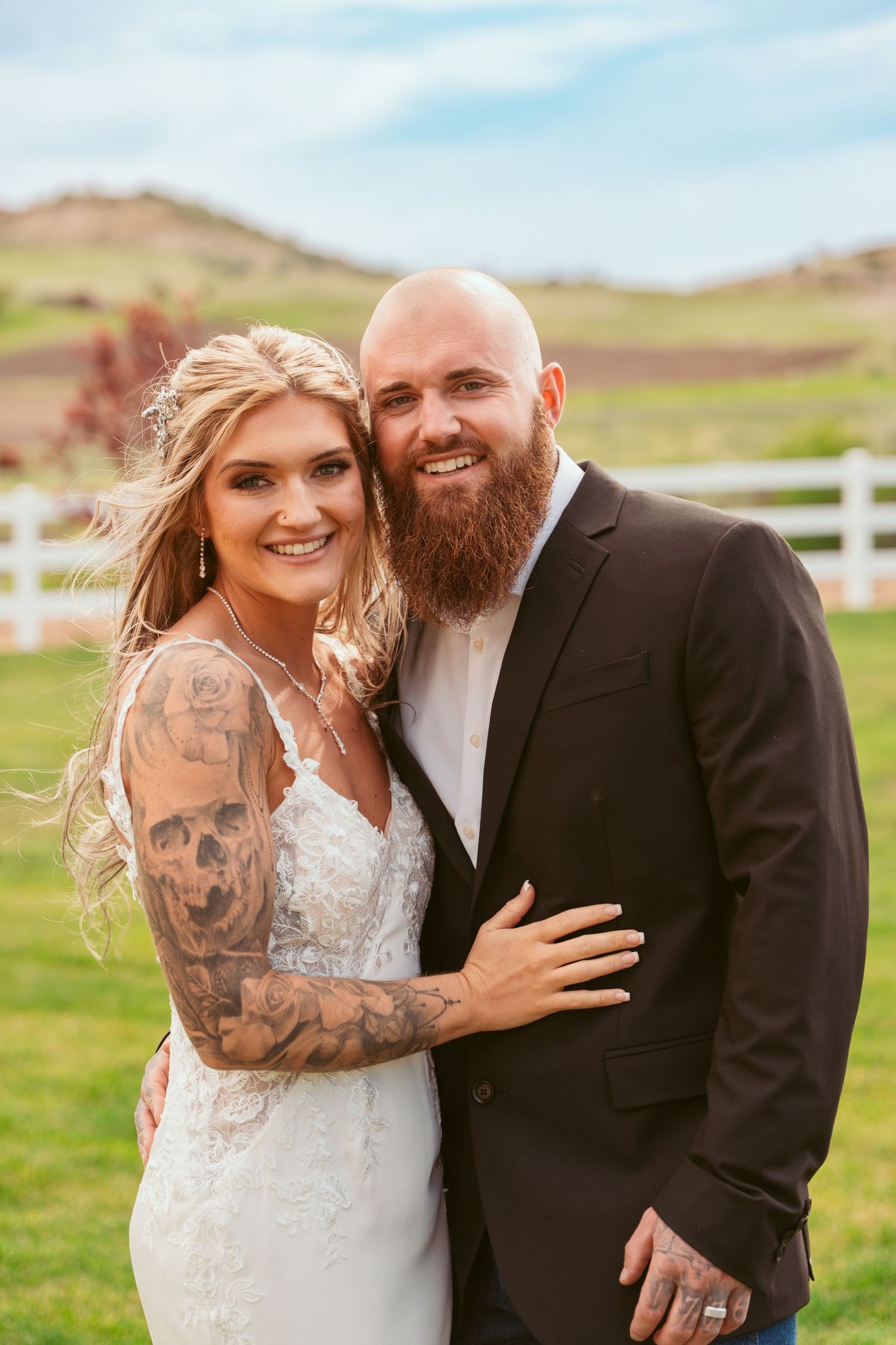 A wedding couple embraces outdoors showing off tattoos and smiles against a scenic mountain backdrop.
