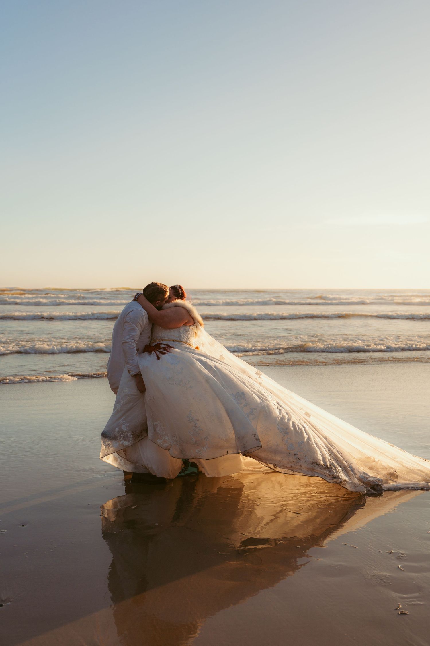 A couple in wedding attire sharing an intimate moment on reflective wet sand at the beach.